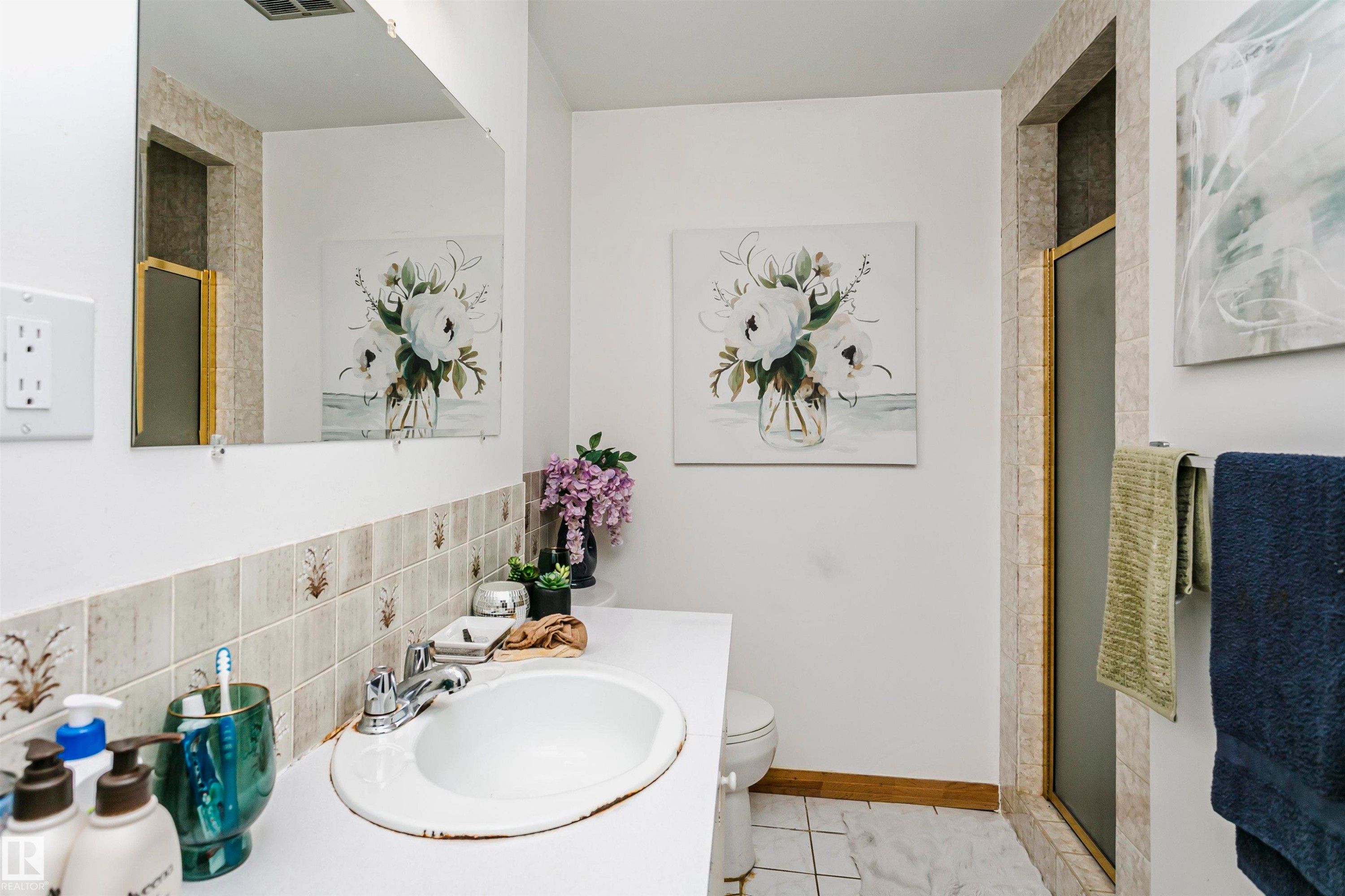 Bathroom featuring a built-in vanity with an integrated sink, a wall-mounted mirror, and a shower enclosure with a gold-tone frame - 14231 80 Street, Edmonton, AB