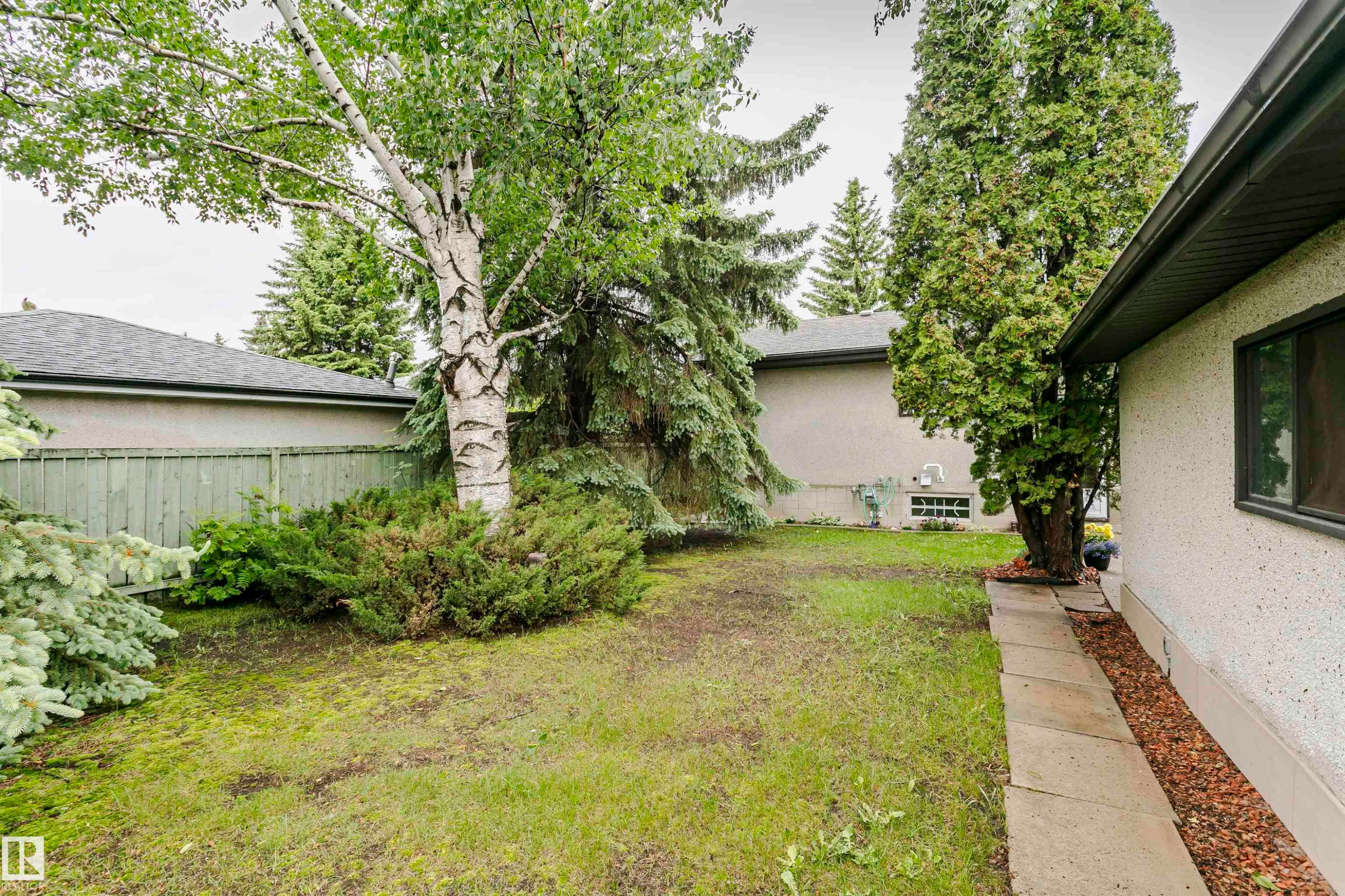 Private backyard space featuring mature trees, established shrubbery, a concrete paver walkway, wood fencing, and a stucco exterior - 14231 80 Street, Edmonton, AB