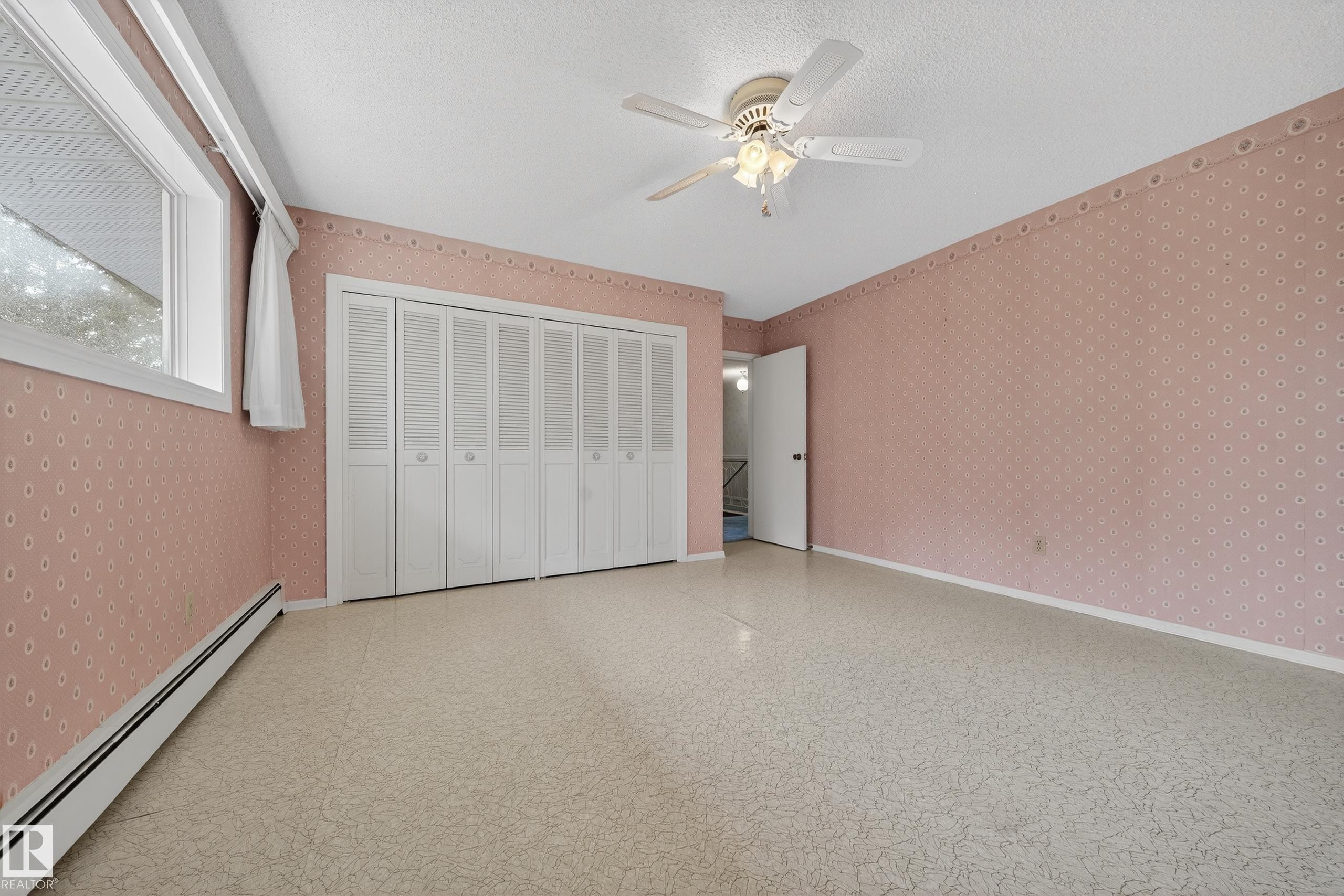 Spacious room with patterned wallpaper, ceiling fan, wide window, baseboard heating, and ample closet space with bi-fold louvered doors - 707 65 Avenue, Edmonton, AB