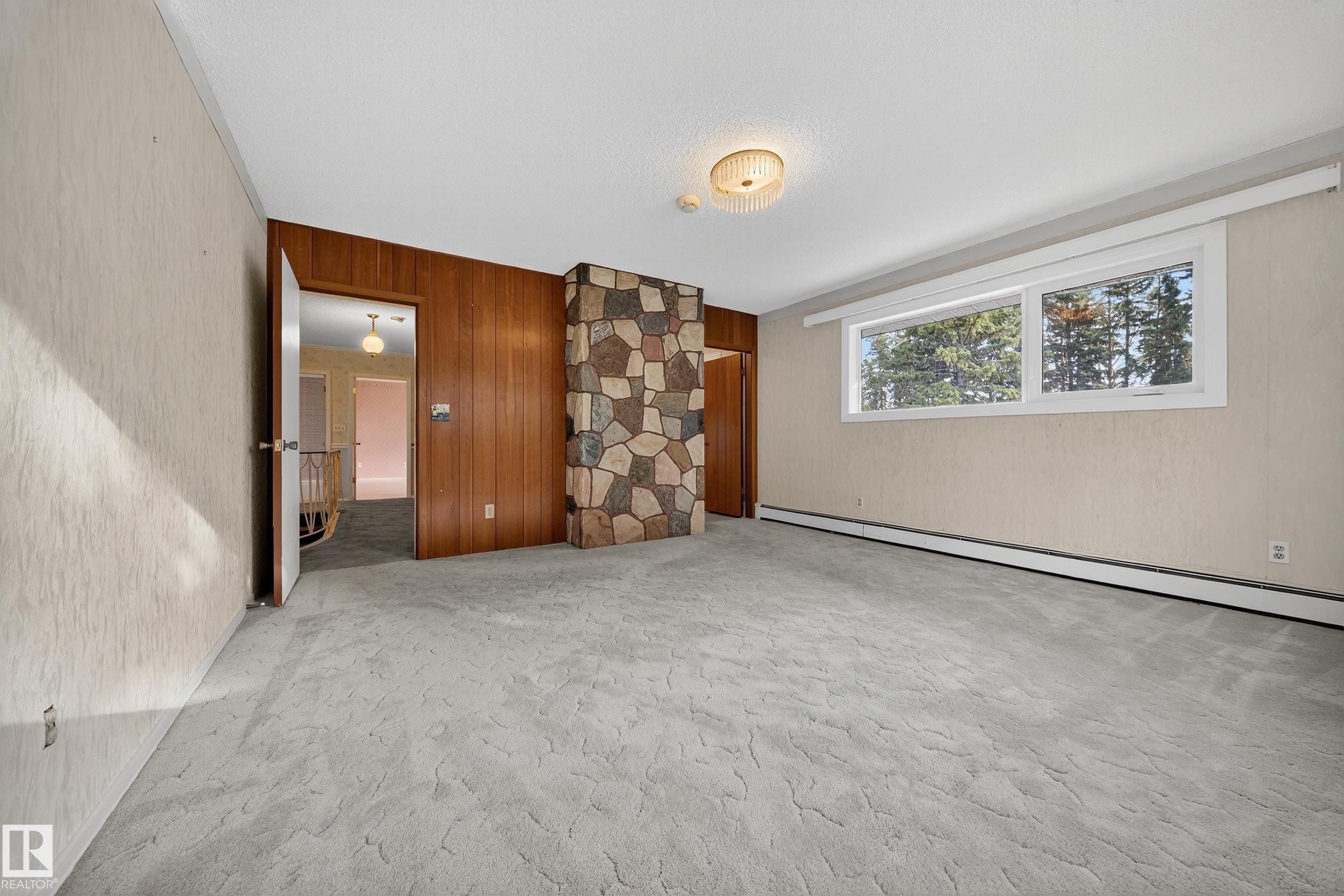 Spacious room featuring light-colored patterned carpeting, a prominent stone-clad architectural element, wood-paneled wall sections, a large window, and baseboard heating - 707 65 Avenue, Edmonton, AB