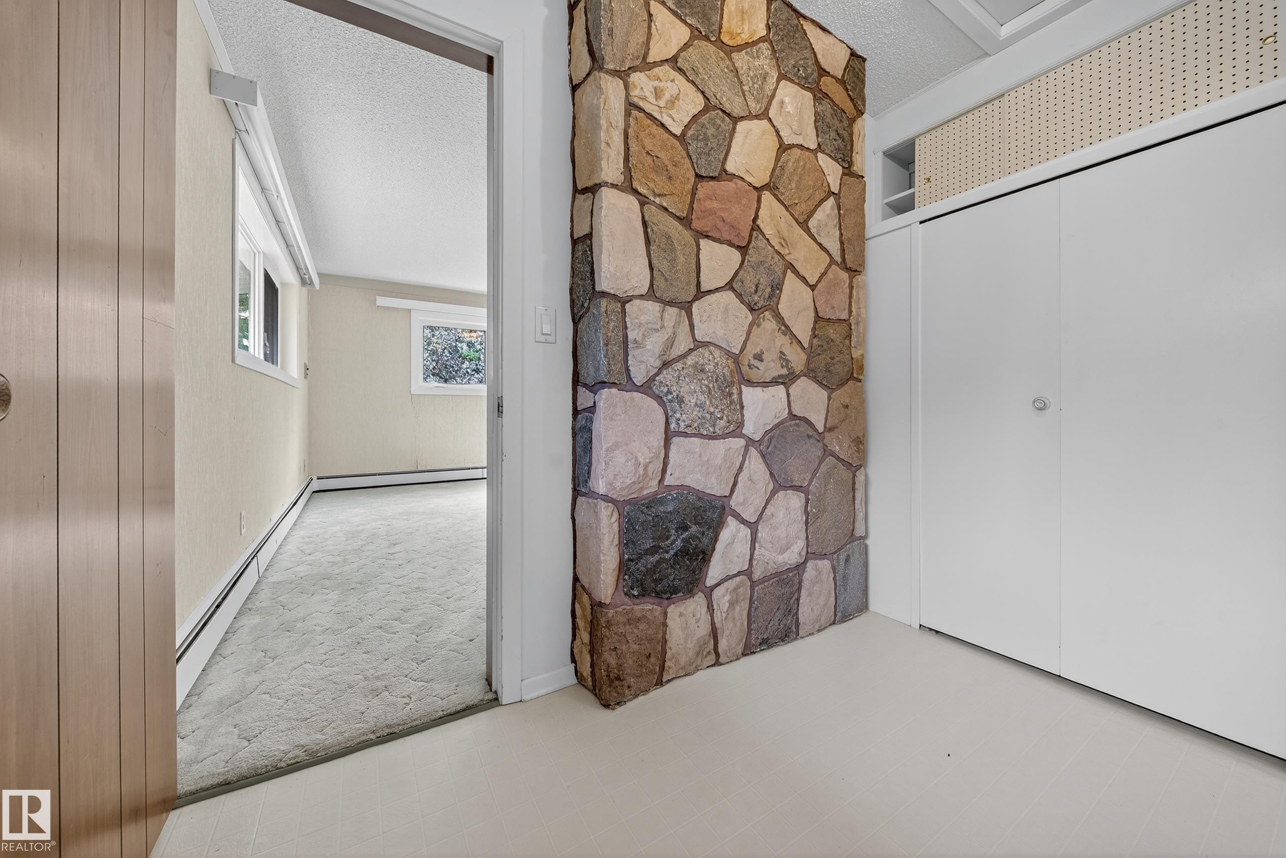 Stone-clad architectural feature with a built-in shelving unit, adjacent to a room with light-colored carpeting, multiple windows, and baseboard heating - 707 65 Avenue, Edmonton, AB