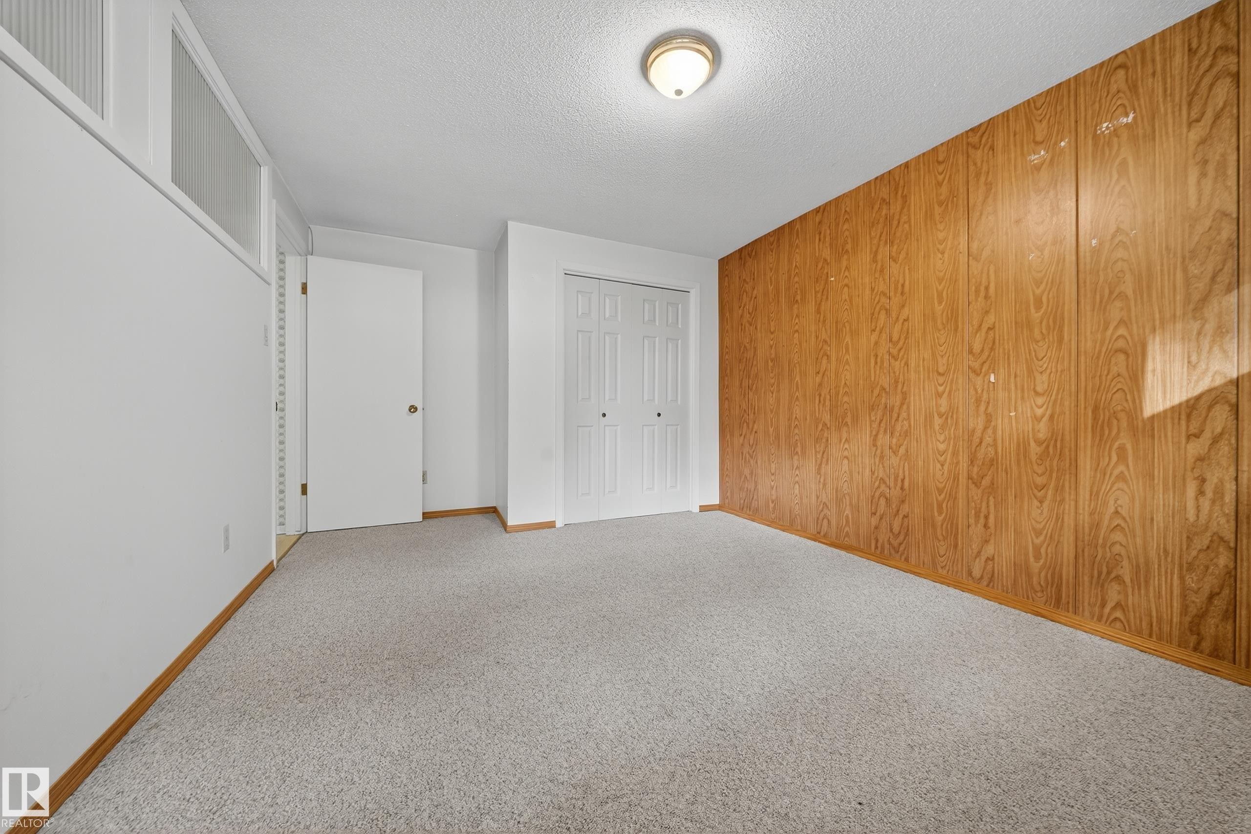 Carpeted room featuring a wood-paneled accent wall, a white painted wall with upper transom windows, a white interior door, and a bi-fold closet - 707 65 Avenue, Edmonton, AB