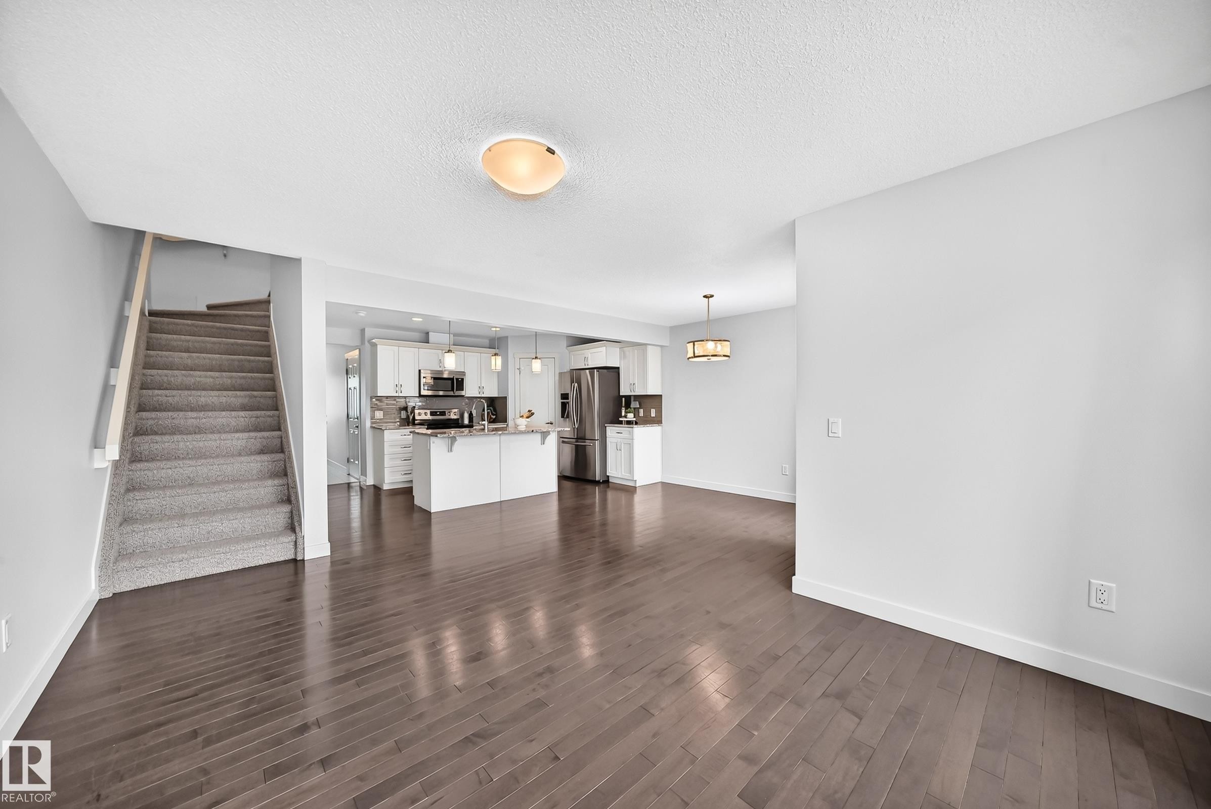 Open-concept living area featuring dark wood-finish flooring, a kitchen with white cabinetry and stainless steel appliances, a kitchen island with seating, a carpeted staircase, and an additional dining area with a decorative chandelier - 2433 Glenridding Boulevard, Edmonton, AB