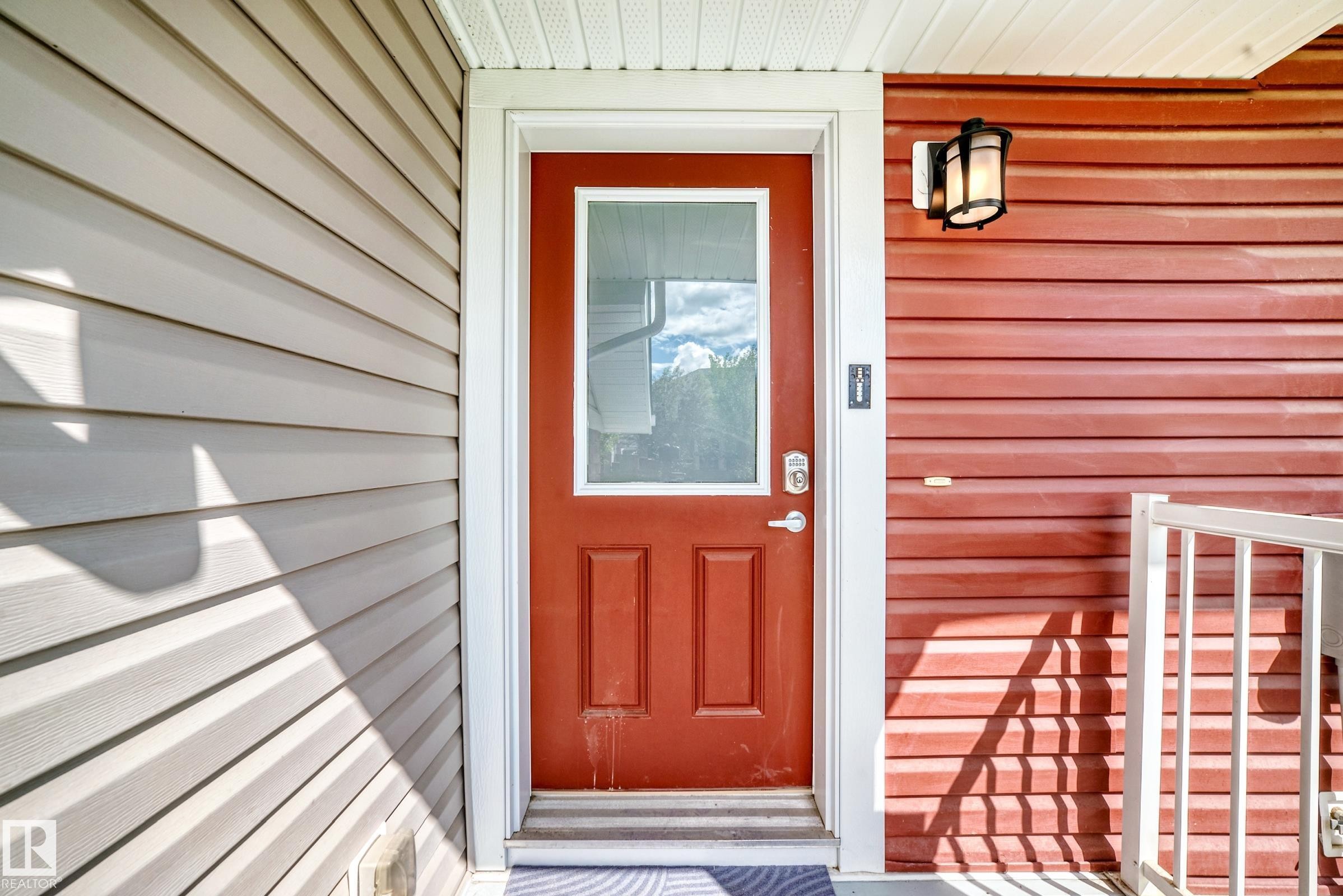 Entryway featuring a red paneled door with an integrated window, contrasting light and dark horizontal siding, white trim, a modern exterior sconce, and a white railing - 2433 Glenridding Boulevard, Edmonton, AB
