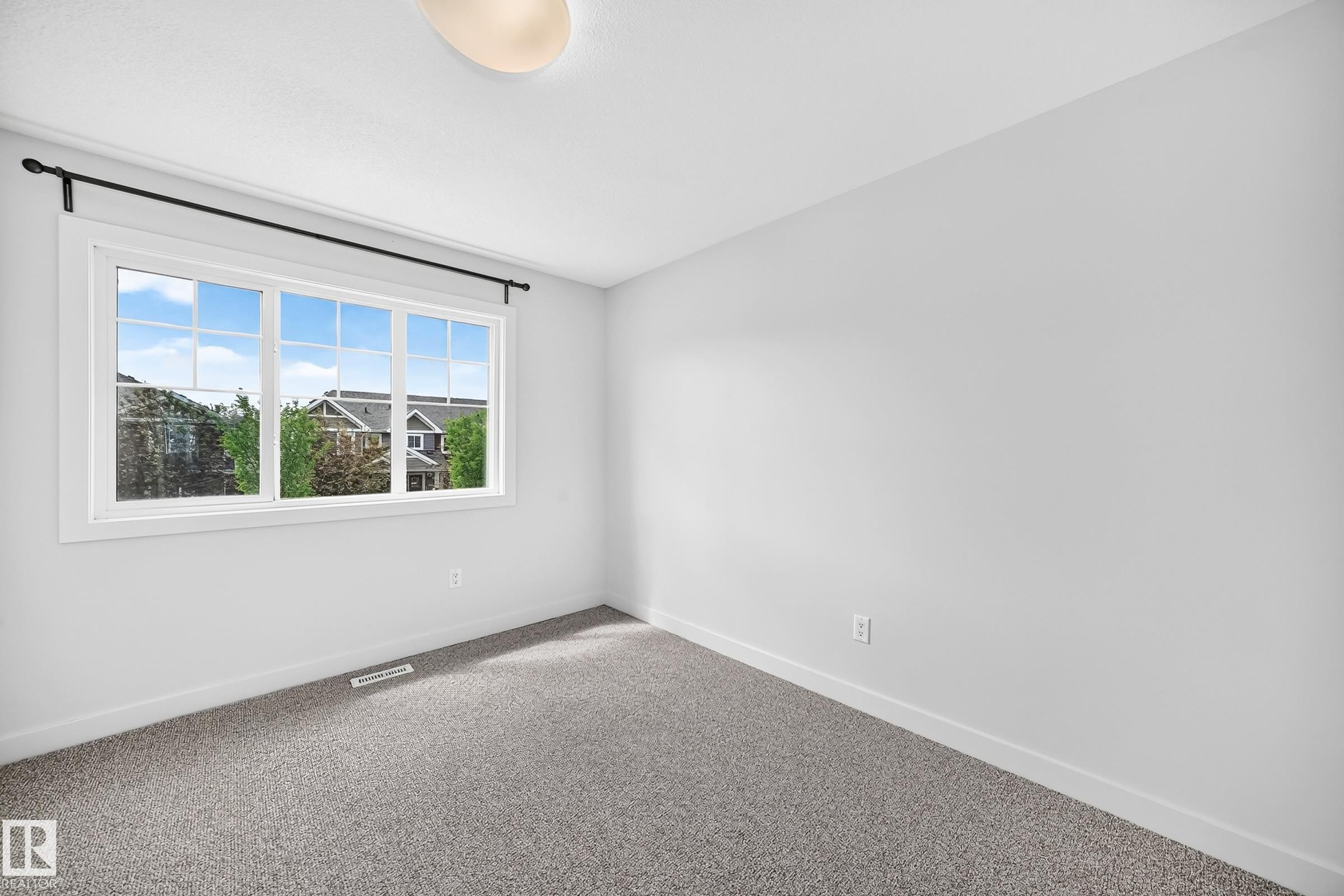 Bright room featuring a large multi-pane window, neutral gray walls, gray carpet flooring, white baseboards, and a flush-mount ceiling light fixture - 2433 Glenridding Boulevard, Edmonton, AB
