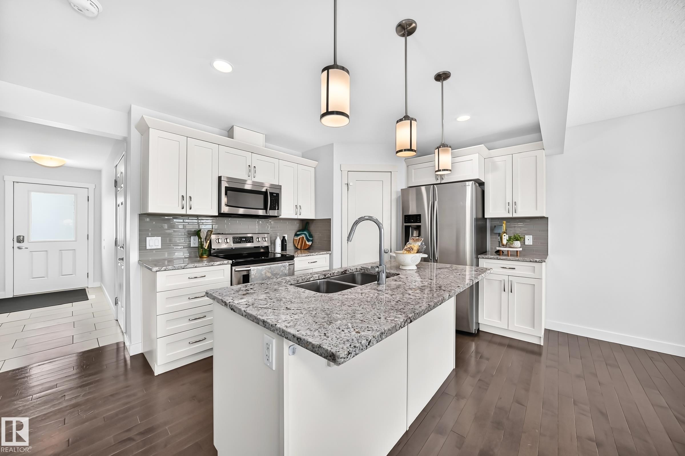 Kitchen featuring a central island with a double basin sink, stainless steel appliances, white cabinetry, a subway tile backsplash, and dark wood-finish flooring - 2433 Glenridding Boulevard, Edmonton, AB
