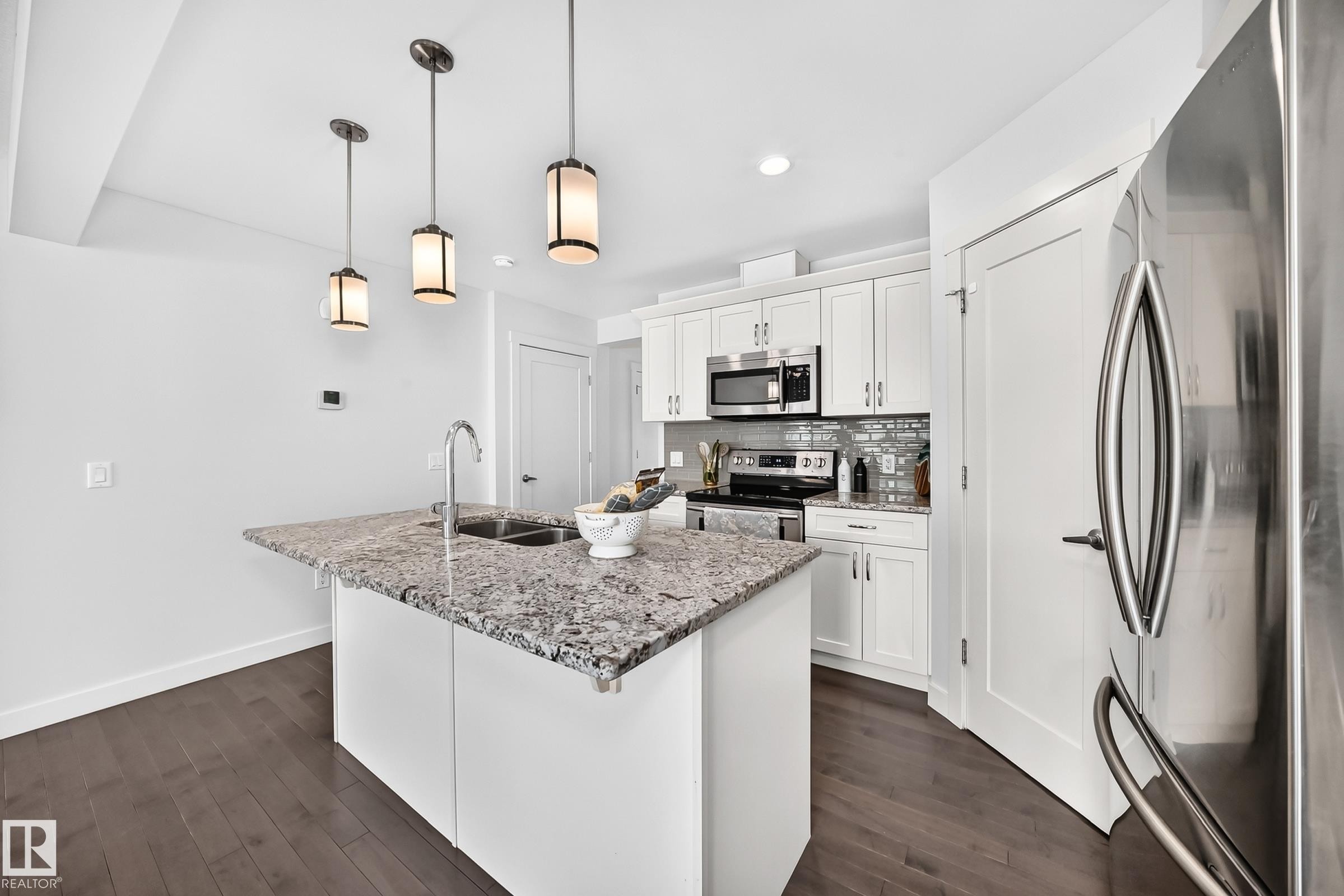 Modern kitchen featuring an island with a double basin sink, three pendant lights, stainless steel appliances, white cabinetry, and wood-finish flooring - 2433 Glenridding Boulevard, Edmonton, AB