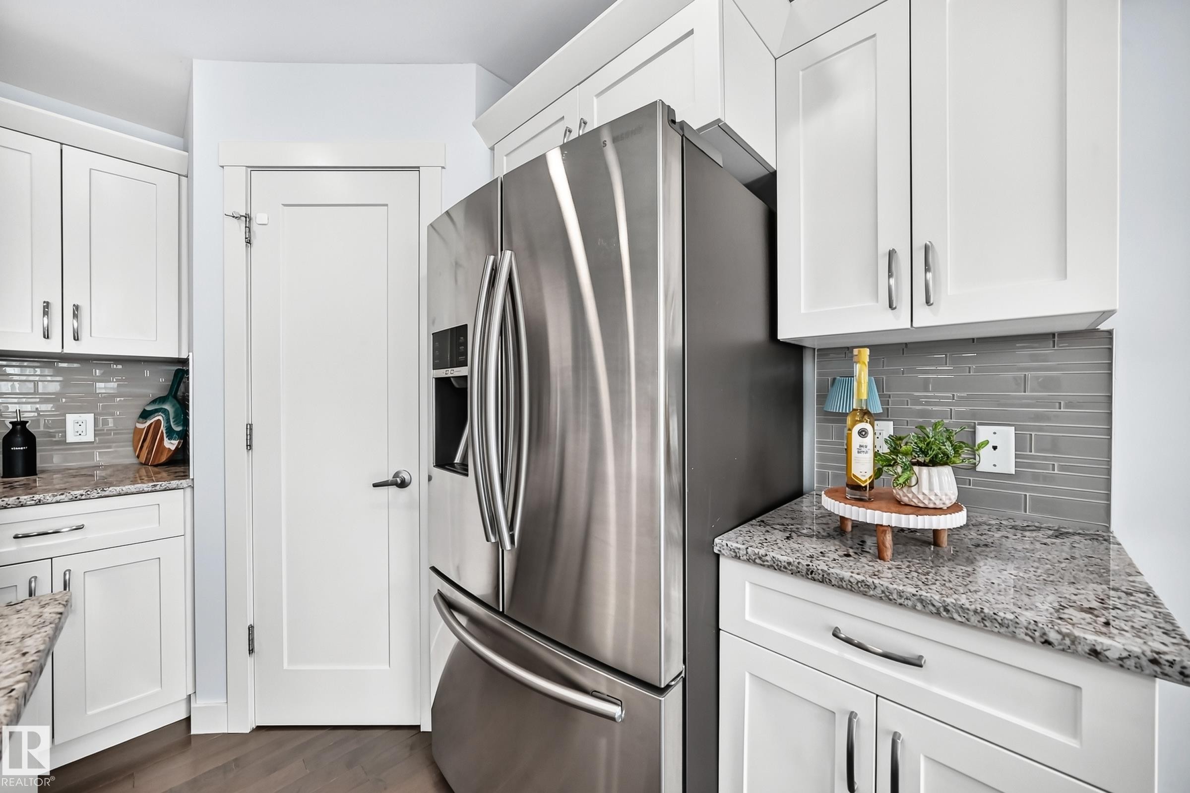 Stainless steel refrigerator within a kitchen featuring white shaker-style cabinetry, light granite countertops, a gray subway tile backsplash, and wood-finish flooring - 2433 Glenridding Boulevard, Edmonton, AB