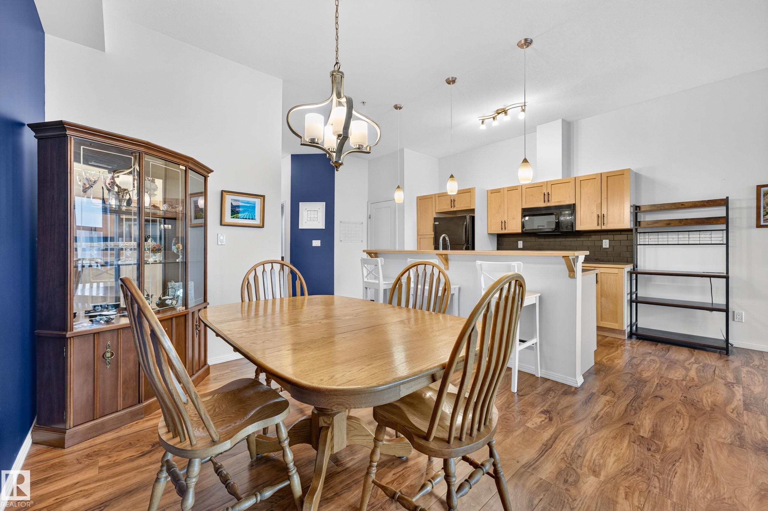 Open-concept dining area featuring wood-finish flooring, a contemporary chandelier, and a kitchen pass-through with pendant lighting - #410 10531 117 St Nw, Edmonton, AB