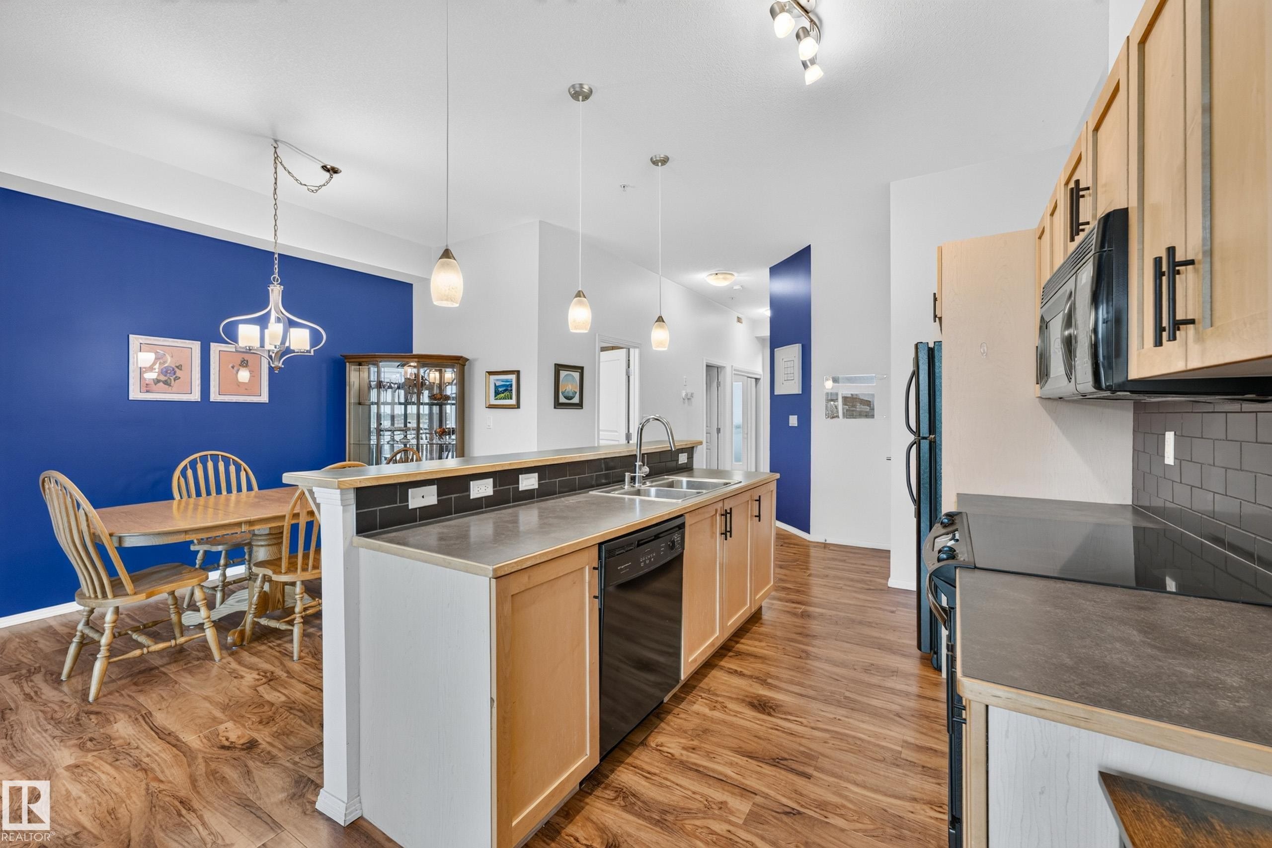 Kitchen and dining area featuring wood-finish flooring, light wood cabinetry, black appliances, a black subway tile backsplash, and a peninsula with integrated sink and seating - #410 10531 117 St Nw, Edmonton, AB