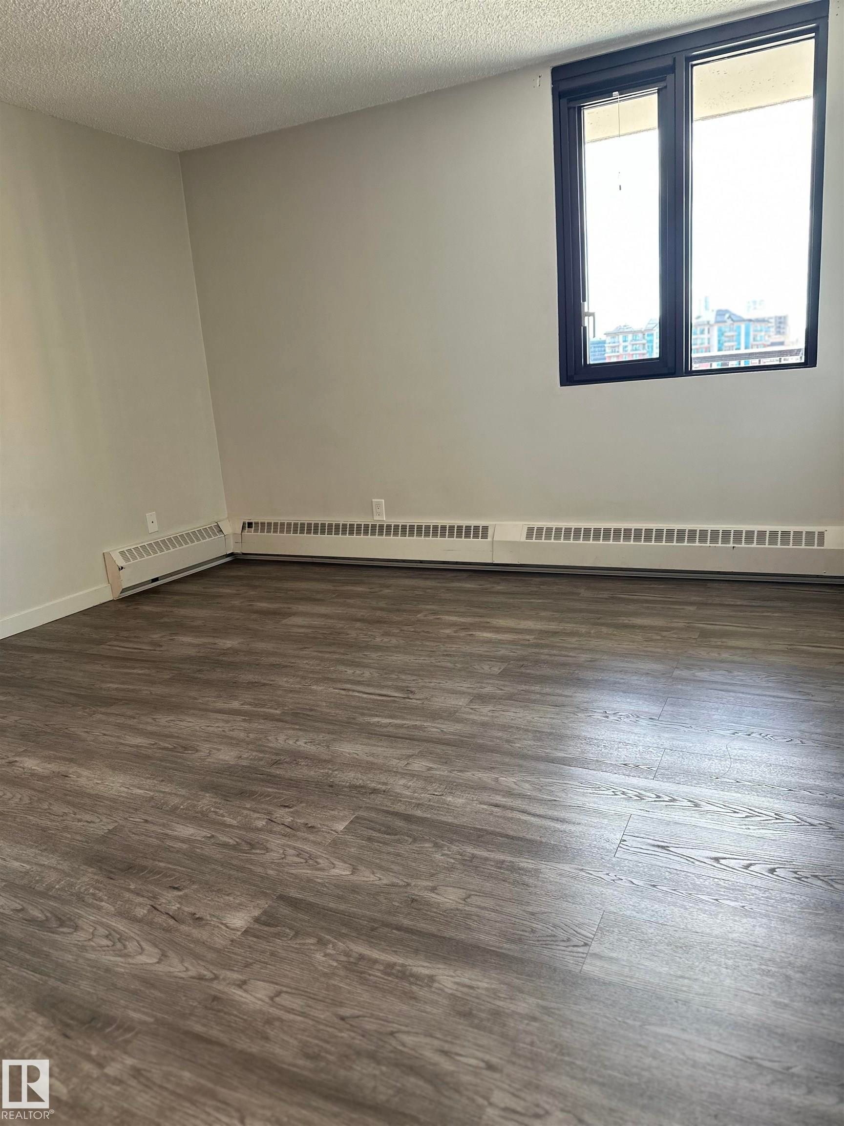 Spacious room with wood-finish flooring, a large window with black trim, and light neutral wall paint - #706 10145 109 St Nw, Edmonton, AB
