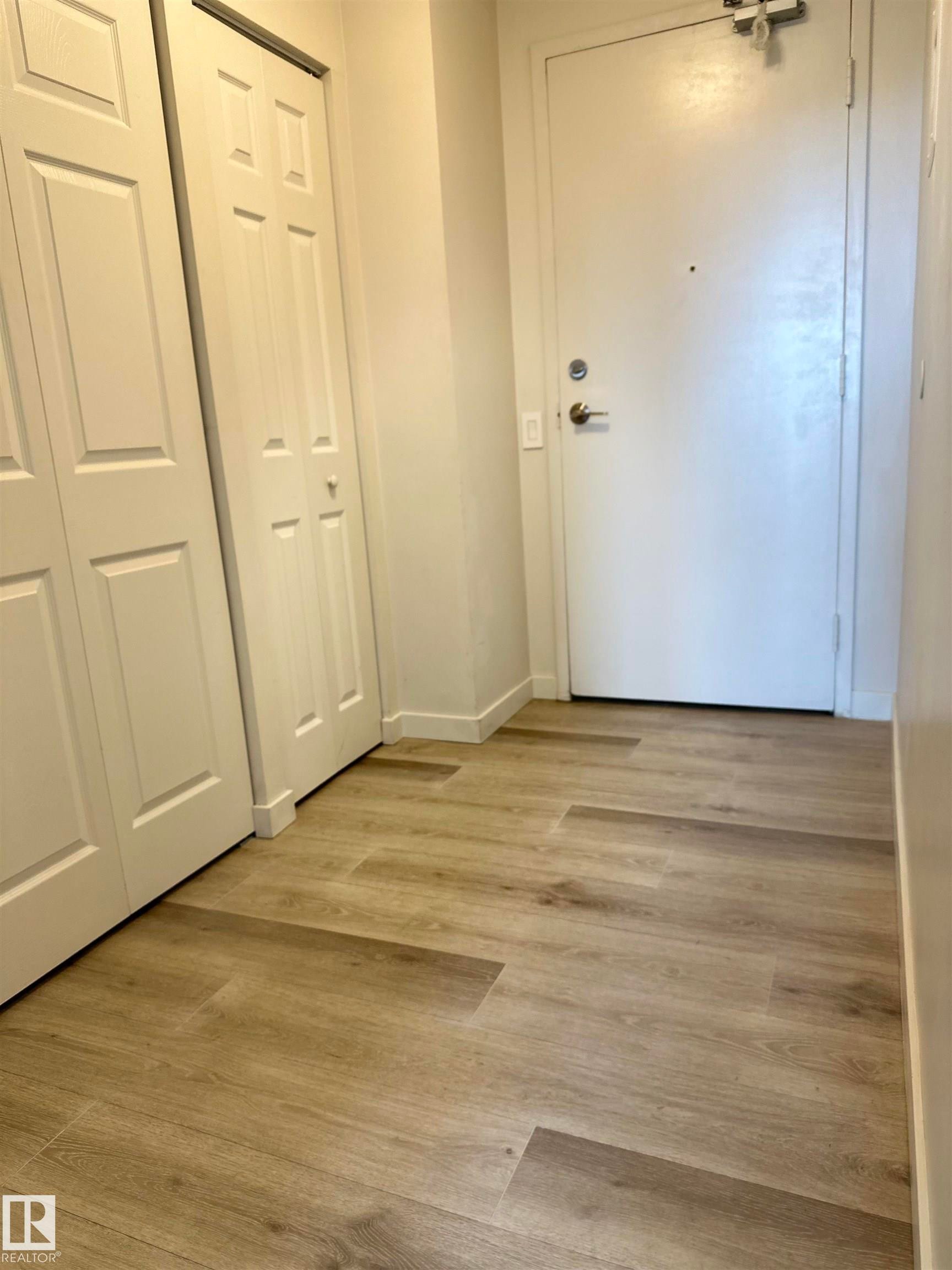 Entryway featuring wood-finish flooring, white paneled bifold doors, and a solid white entry door with brass hardware - #706 10145 109 St Nw, Edmonton, AB