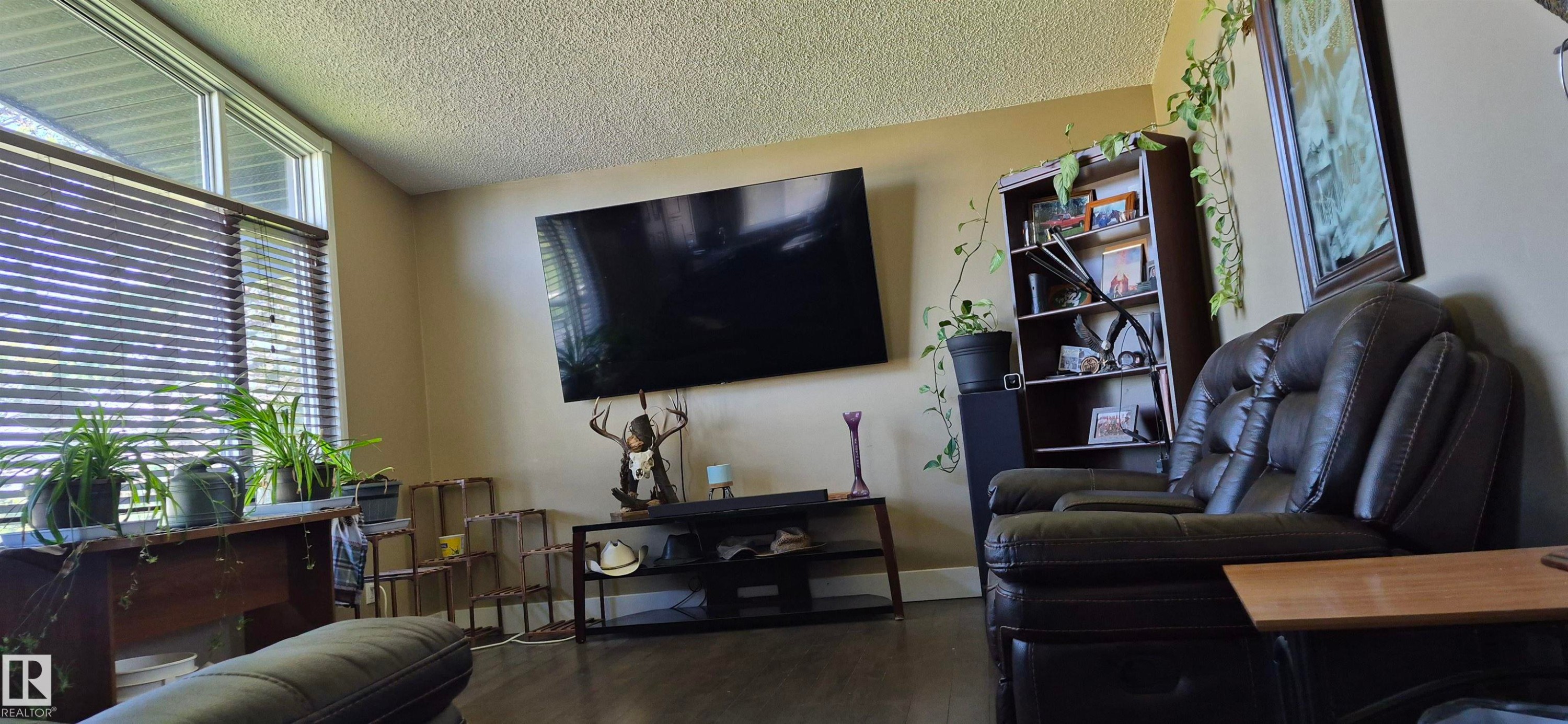 Spacious living area featuring wood-finish flooring, neutral wall tones, and a prominent window with horizontal blinds - 10523 105 Street, Westlock, AB