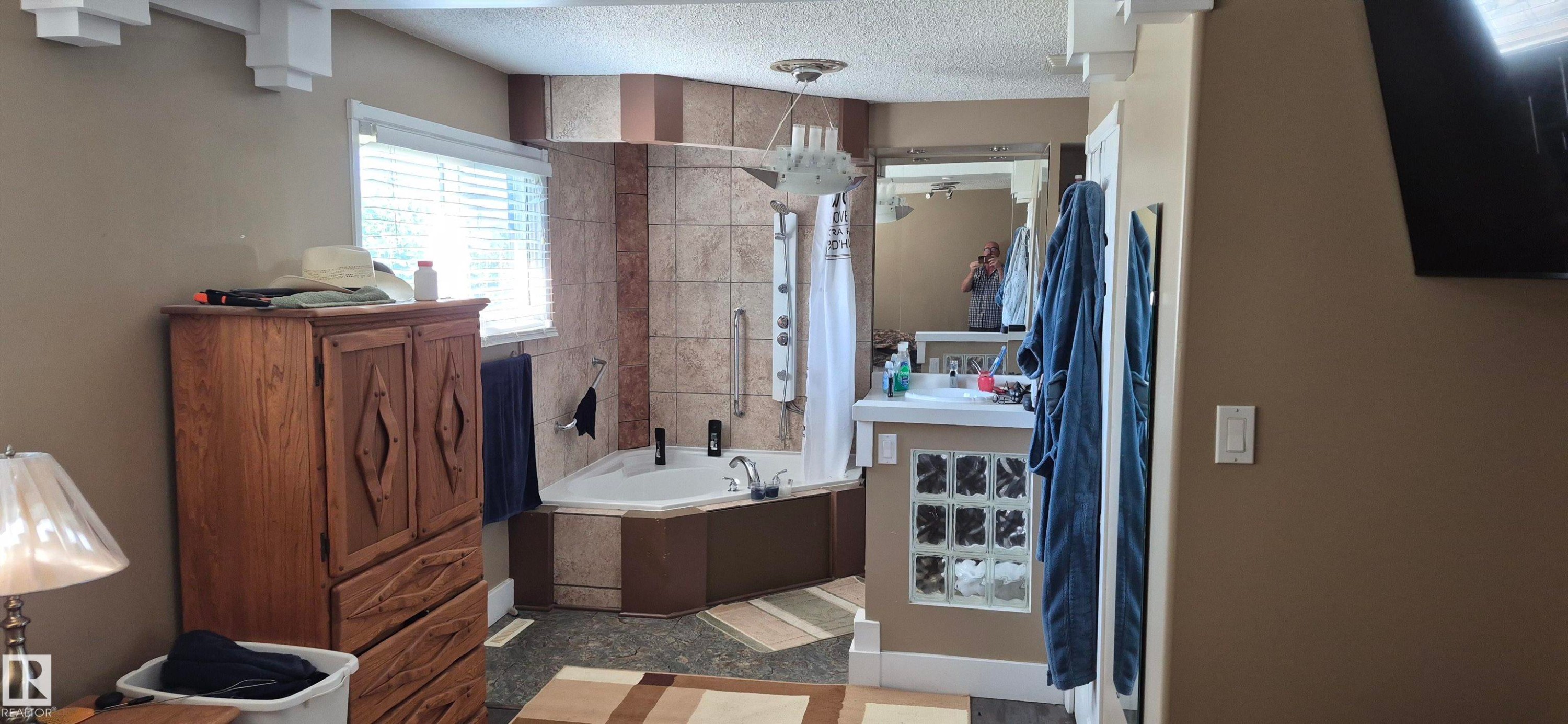 Corner jetted tub with tiled surround, adjacent to a single vanity featuring a rectangular sink, glass block detailing, and a mirrored wall - 10523 105 Street, Westlock, AB