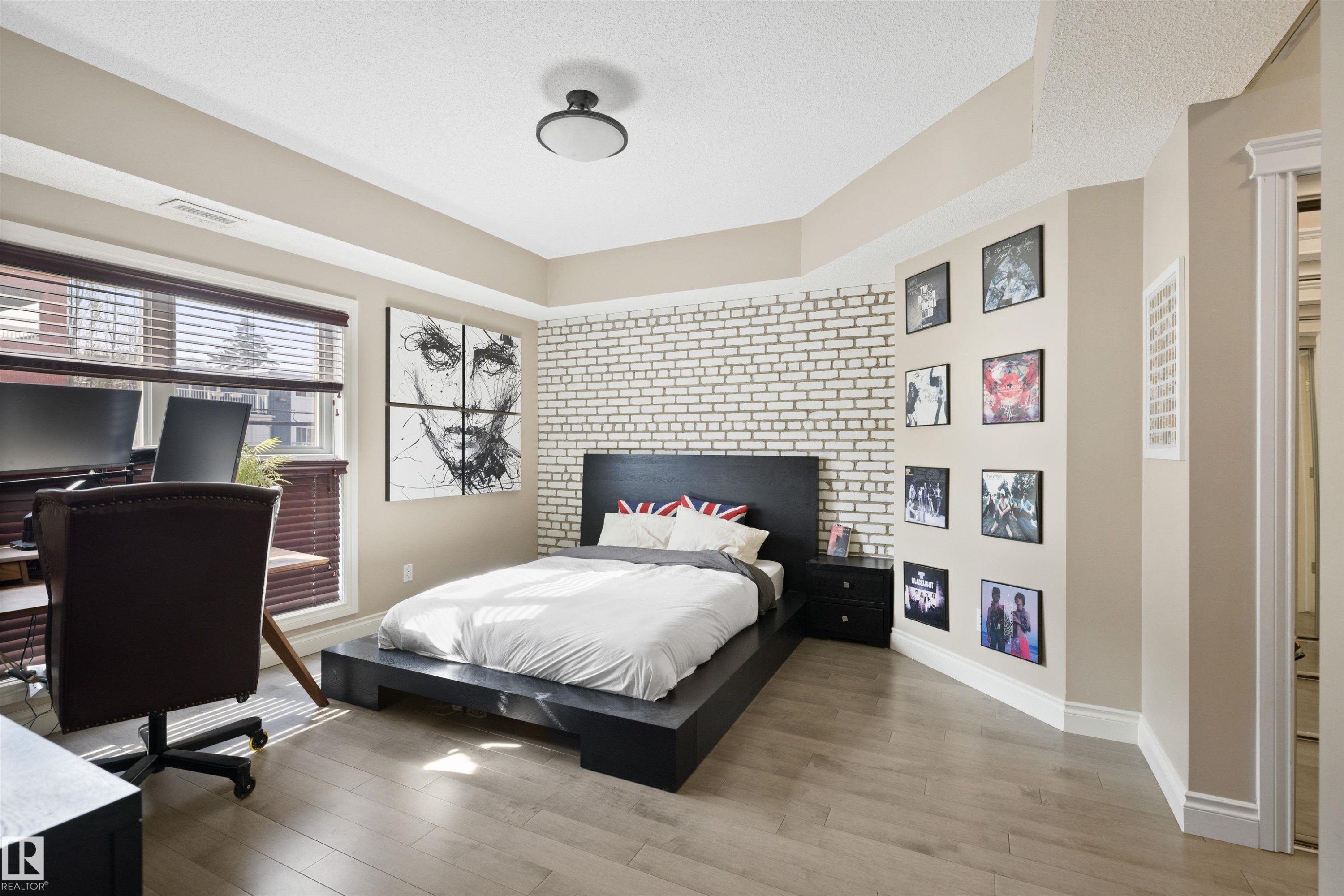 Spacious room featuring light wood-finish flooring, a prominent white brick accent wall, and a large window with horizontal blinds - #206 10232 115 St Nw, Edmonton, AB
