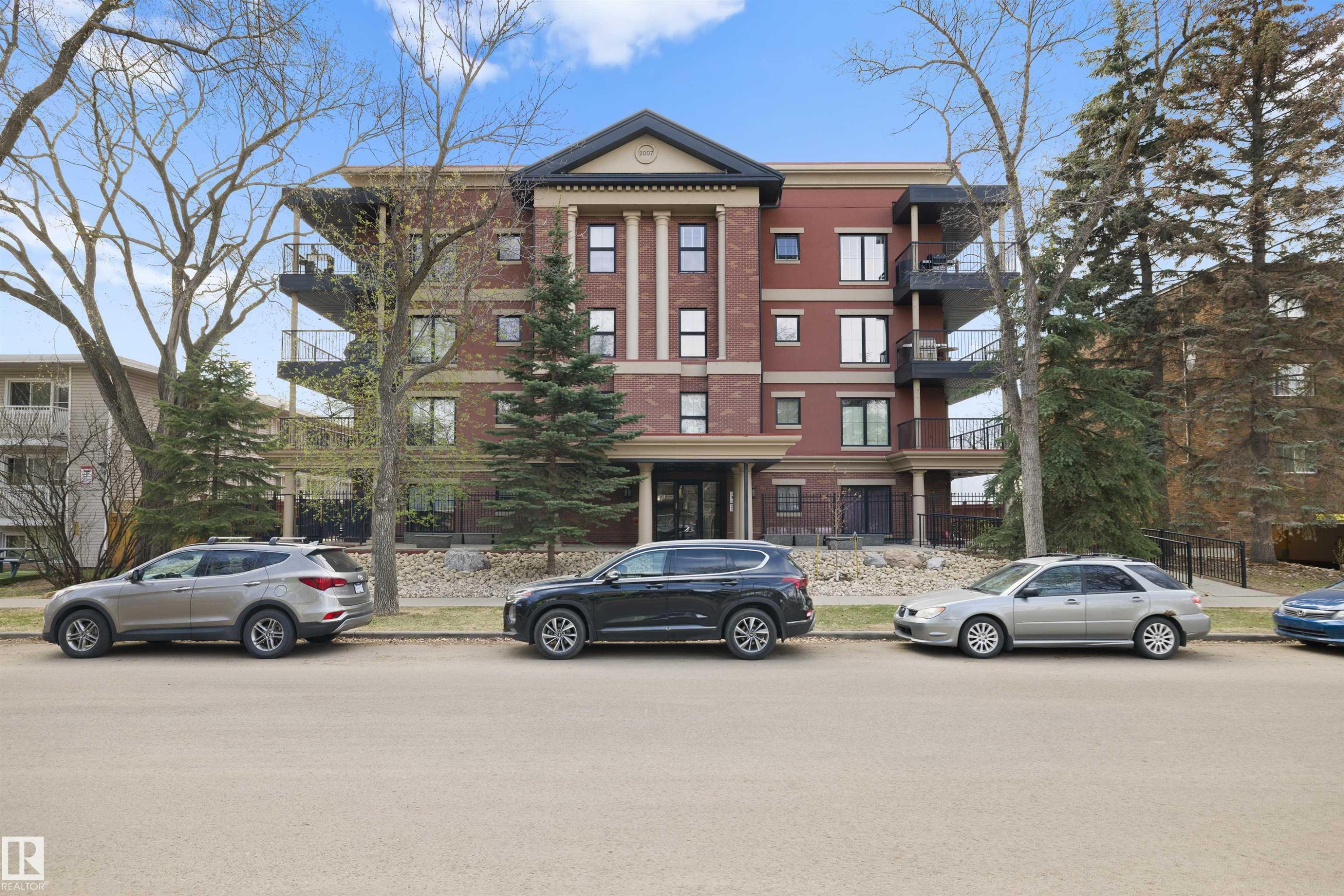 Multi-story residential building featuring brick facade, prominent architectural columns, multiple balconies, and mature landscaping - #206 10232 115 St Nw, Edmonton, AB