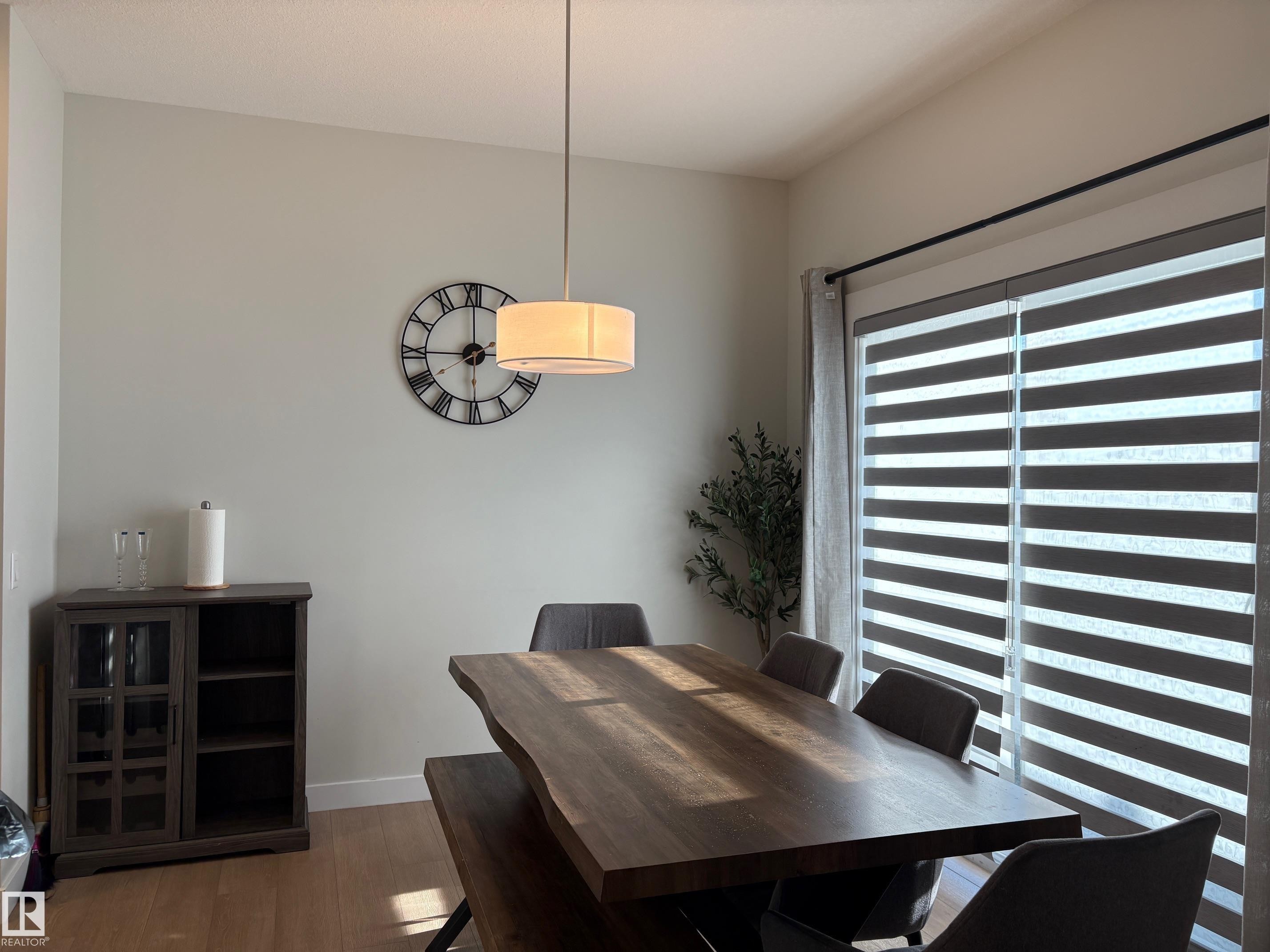 Dining area featuring wood-finish flooring, a drum pendant light fixture, and a large window with horizontal layered blinds - 9952 227 Street, Edmonton, AB