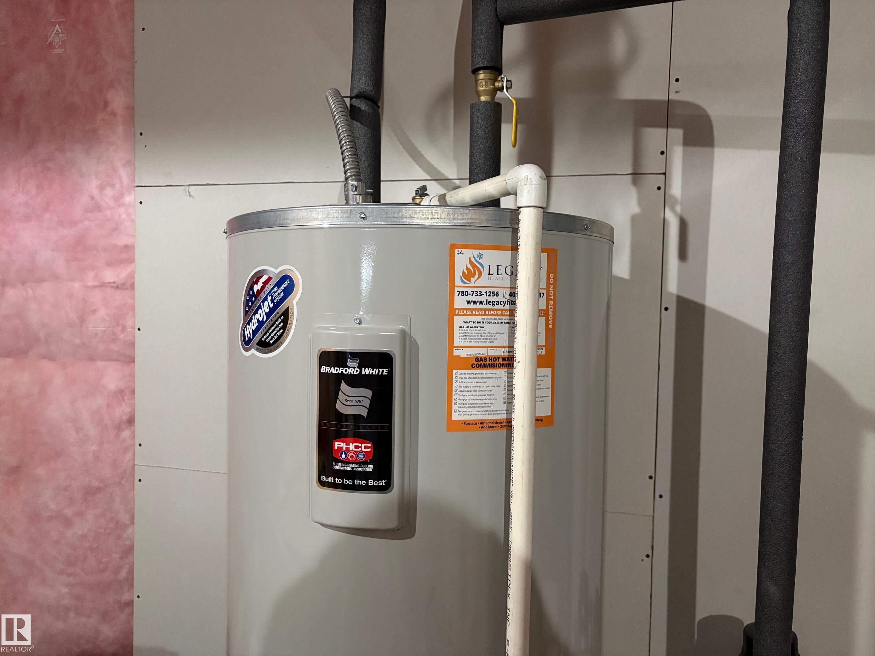 Bradford White water heater unit with insulated piping and a brass shut-off valve, positioned against drywall and visible pink insulation - 9952 227 Street, Edmonton, AB