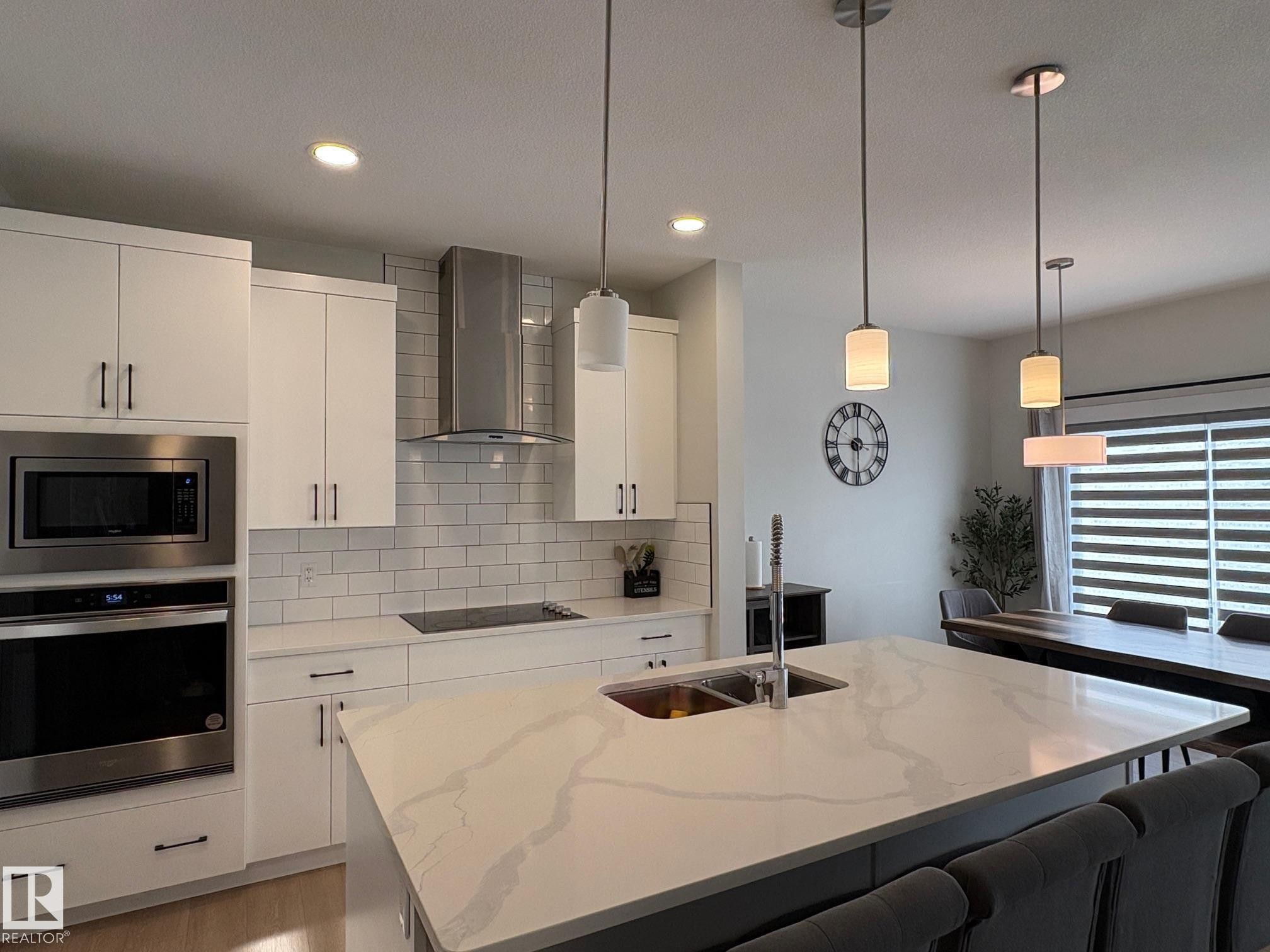 Gourmet kitchen featuring a large central island with double basin sink, white cabinetry with dark hardware, integrated stainless steel appliances, and a white subway tile backsplash - 9952 227 Street, Edmonton, AB