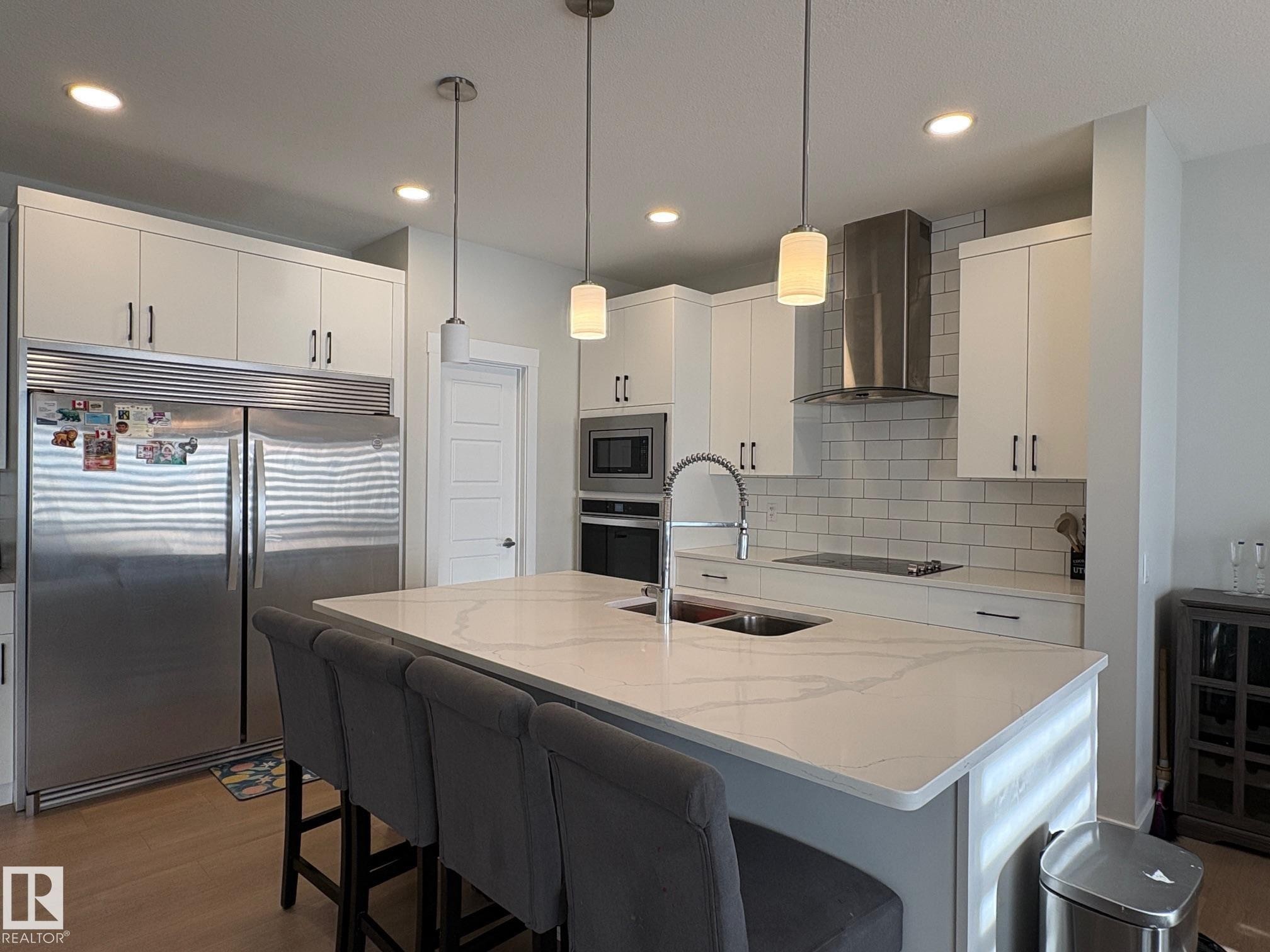 Modern kitchen featuring a large center island with integrated sink, white cabinetry with dark hardware, stainless steel appliances, and subway tile backsplash - 9952 227 Street, Edmonton, AB
