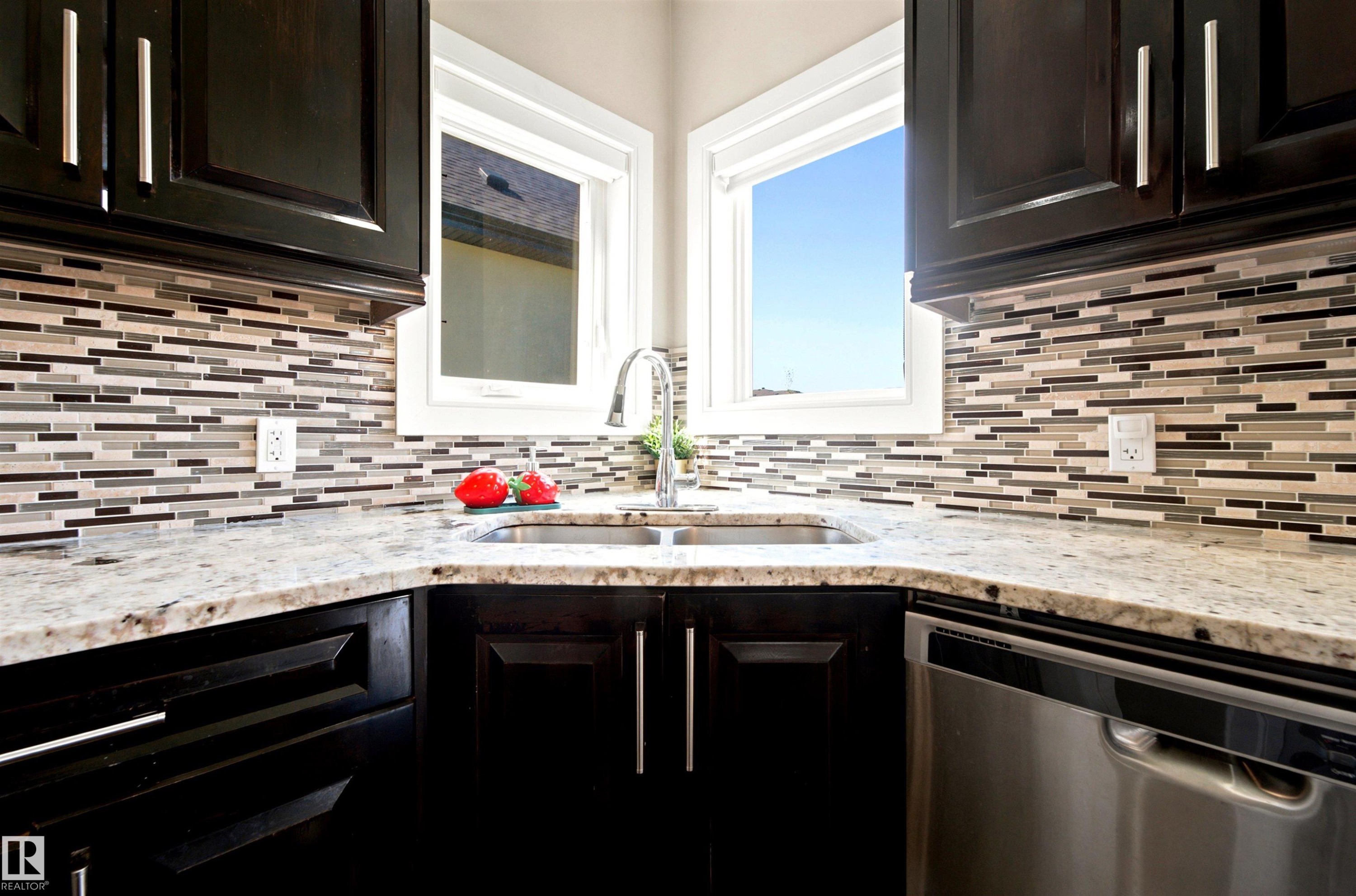 Kitchen corner featuring dark wood-finish cabinetry with brushed nickel hardware, light-toned stone countertops, and a full-height mosaic tile backsplash - 5110 Rue Eaglemont, Beaumont, AB