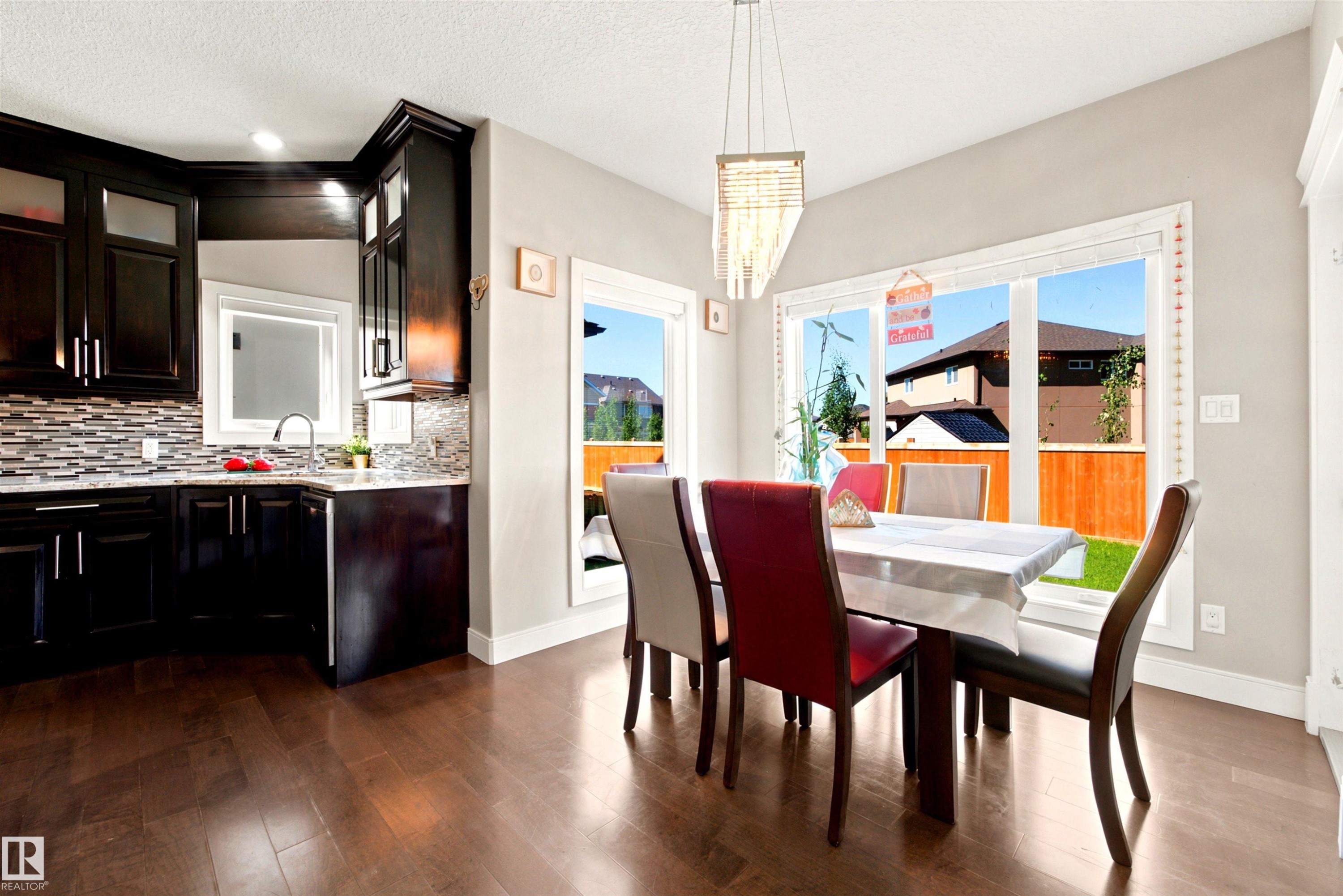 Open-concept dining area with wood-finish flooring, a contemporary linear pendant light fixture, and large windows providing abundant natural light - 5110 Rue Eaglemont, Beaumont, AB