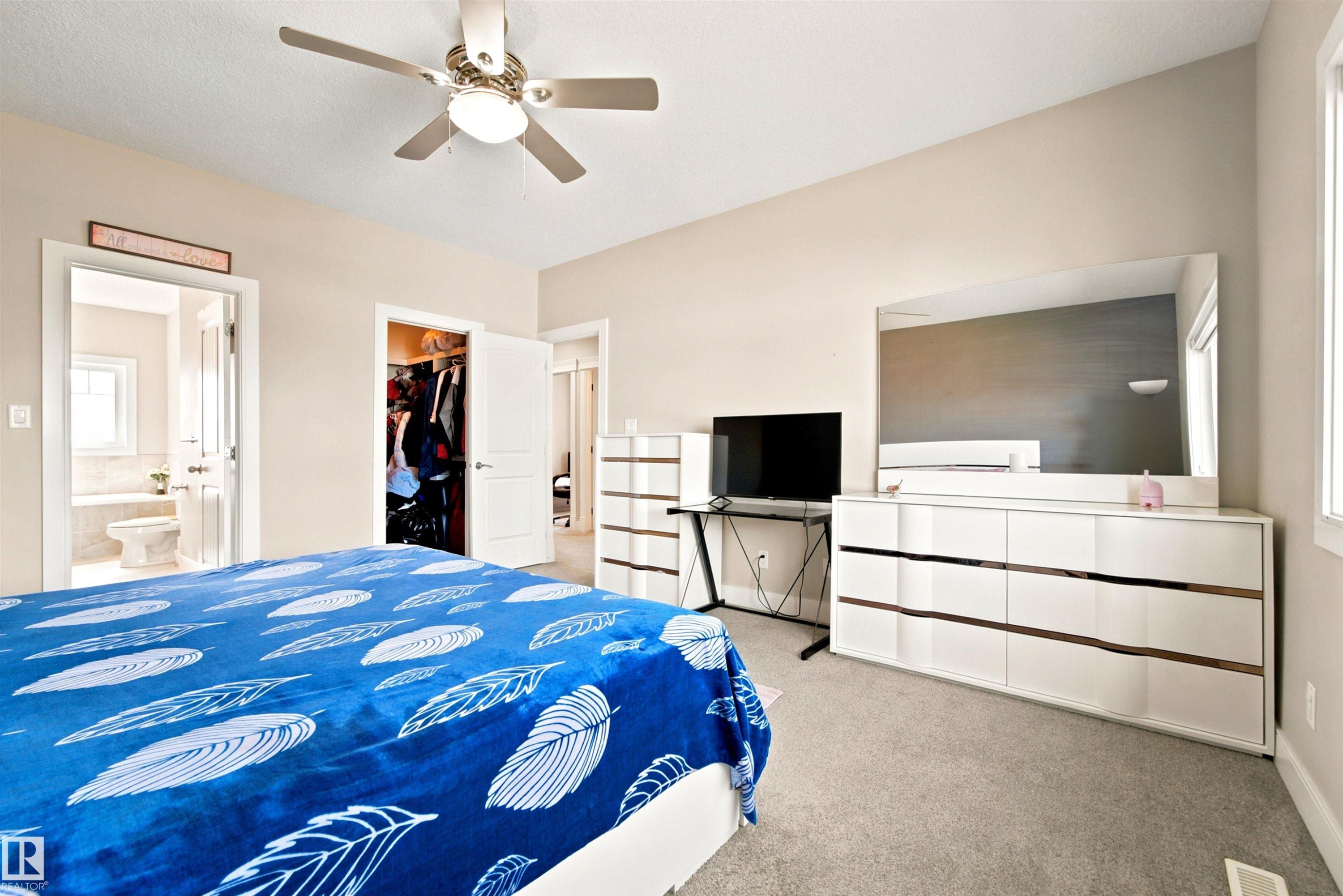 Carpeted room featuring a ceiling fan with integrated lighting, a walk-in closet, a bathroom entry, and a window providing natural light - 5110 Rue Eaglemont, Beaumont, AB
