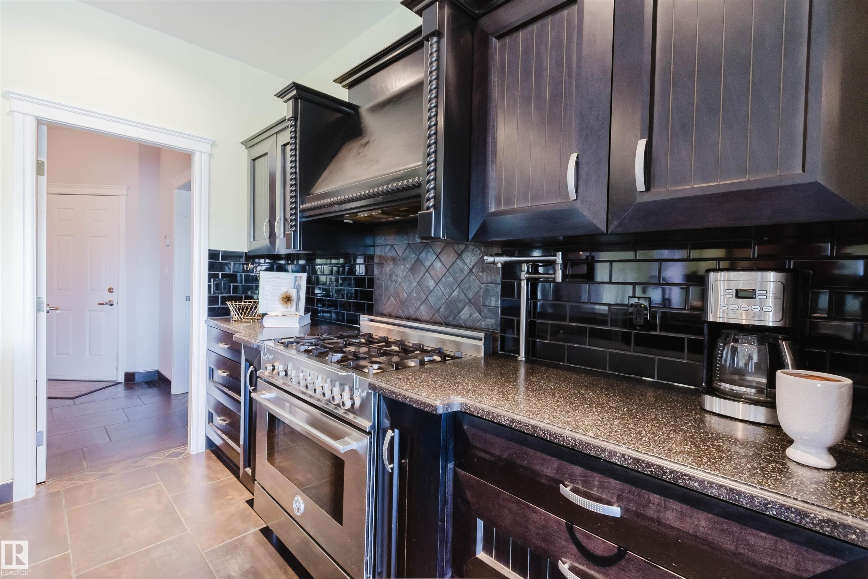 Kitchen featuring dark wood-finish cabinetry, speckled countertops, a stainless steel range with oven, and a black subway tile backsplash with a diamond pattern accent - 3438 West Landing Landing, Edmonton, AB