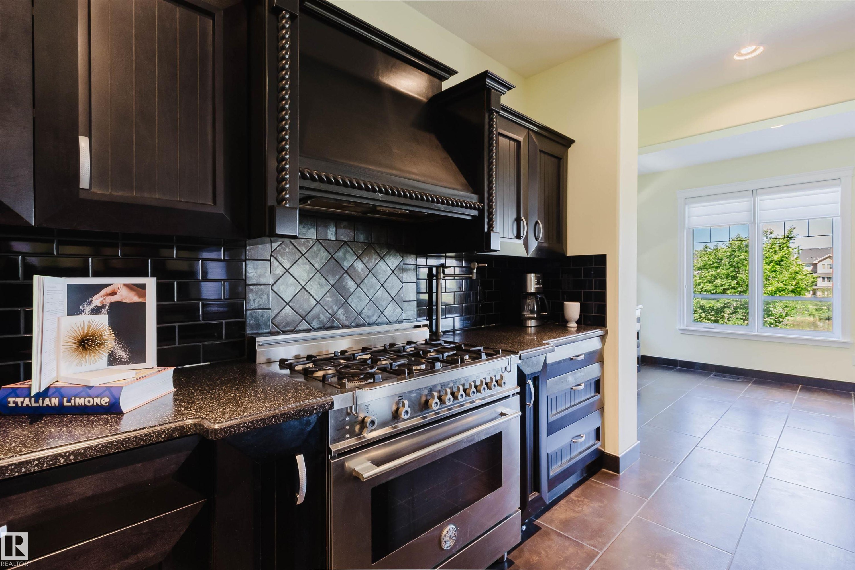 Gourmet kitchen featuring dark wood cabinetry, a stainless steel range with a custom hood, black speckled countertops, and a black subway tile backsplash with a decorative inset - 3438 West Landing Landing, Edmonton, AB