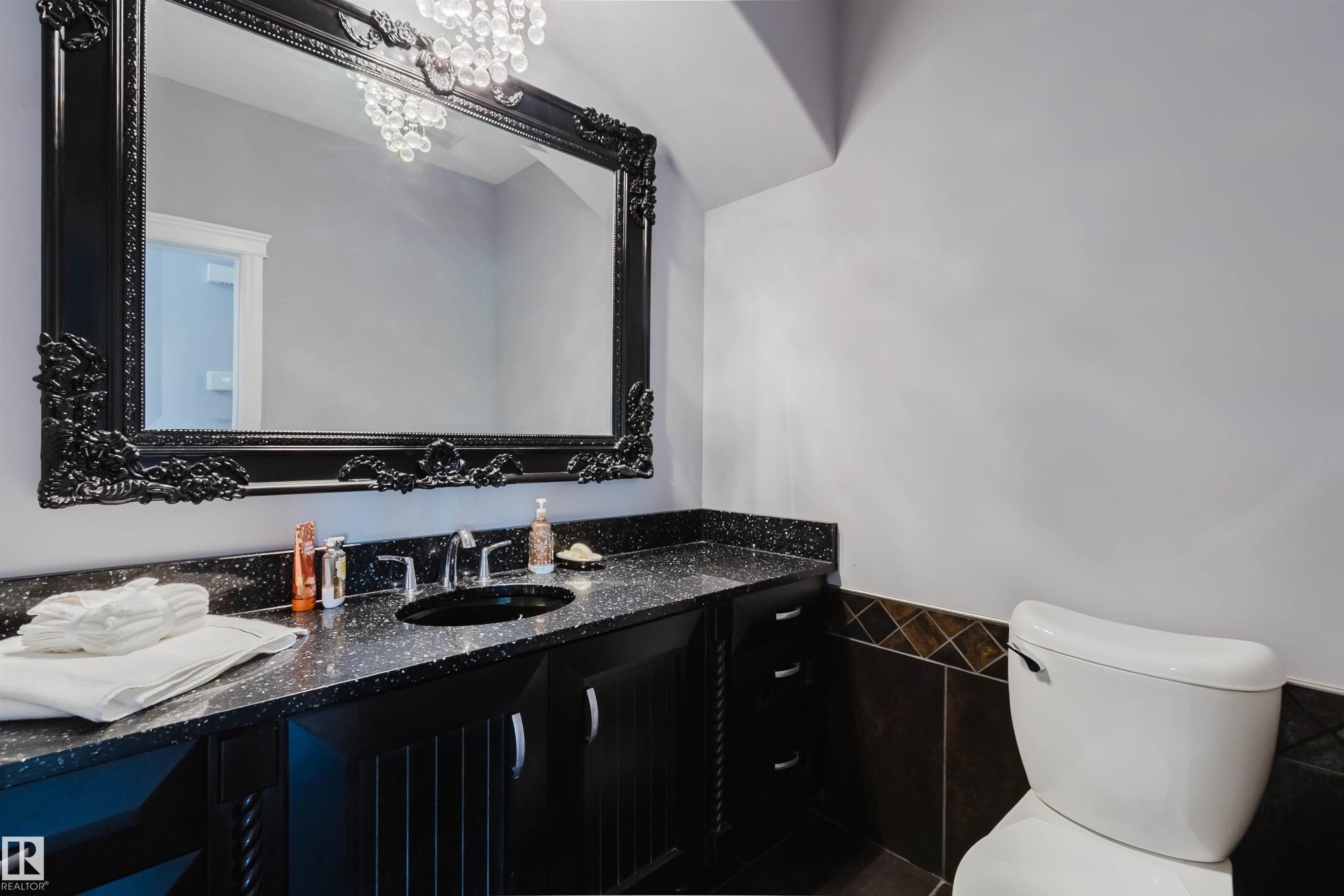 Bathroom featuring a large ornate black framed mirror, a black countertop with integrated sink, black cabinetry, a crystal chandelier, and wall tiling - 3438 West Landing Landing, Edmonton, AB