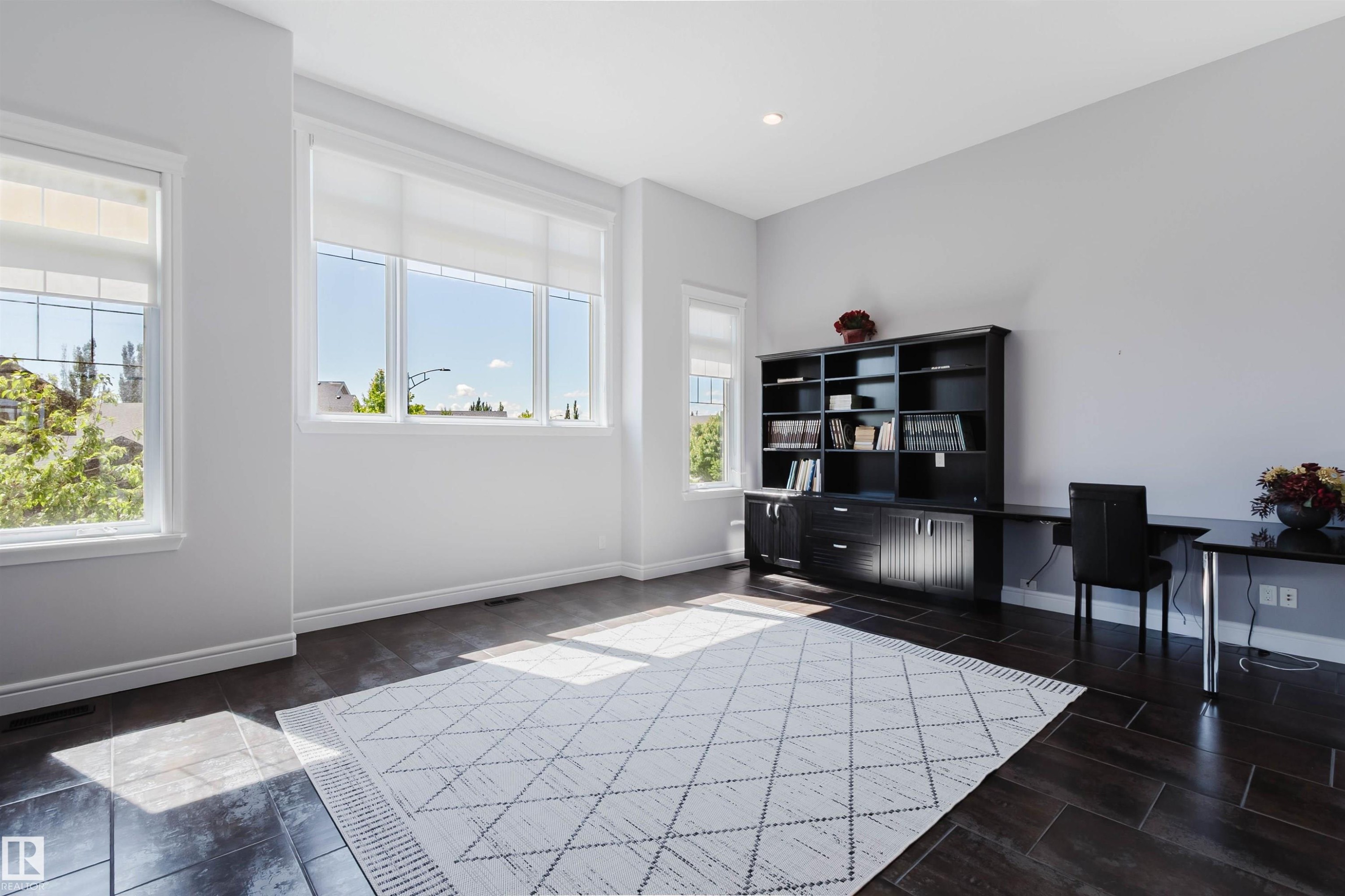 Spacious room featuring dark wood-finish flooring, a built-in dark wood bookcase with integrated desk, multiple large windows, recessed lighting, and crisp white baseboards - 3438 West Landing Landing, Edmonton, AB