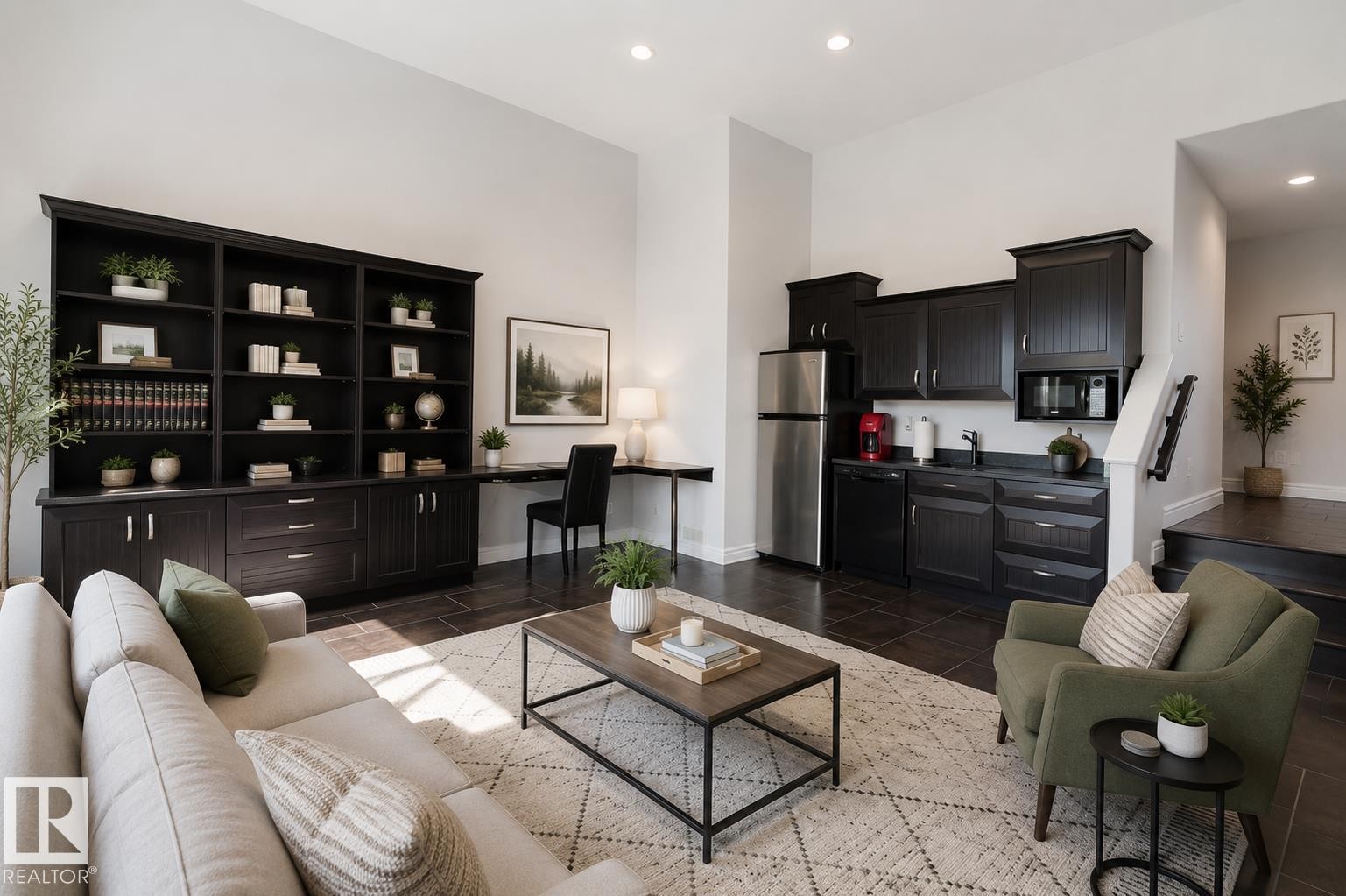 Open-concept living area featuring a built-in dark wood-finish shelving unit, integrated desk space, and a compact kitchen area with dark cabinetry, stainless steel appliances, and a built-in microwave - 3438 West Landing Landing, Edmonton, AB