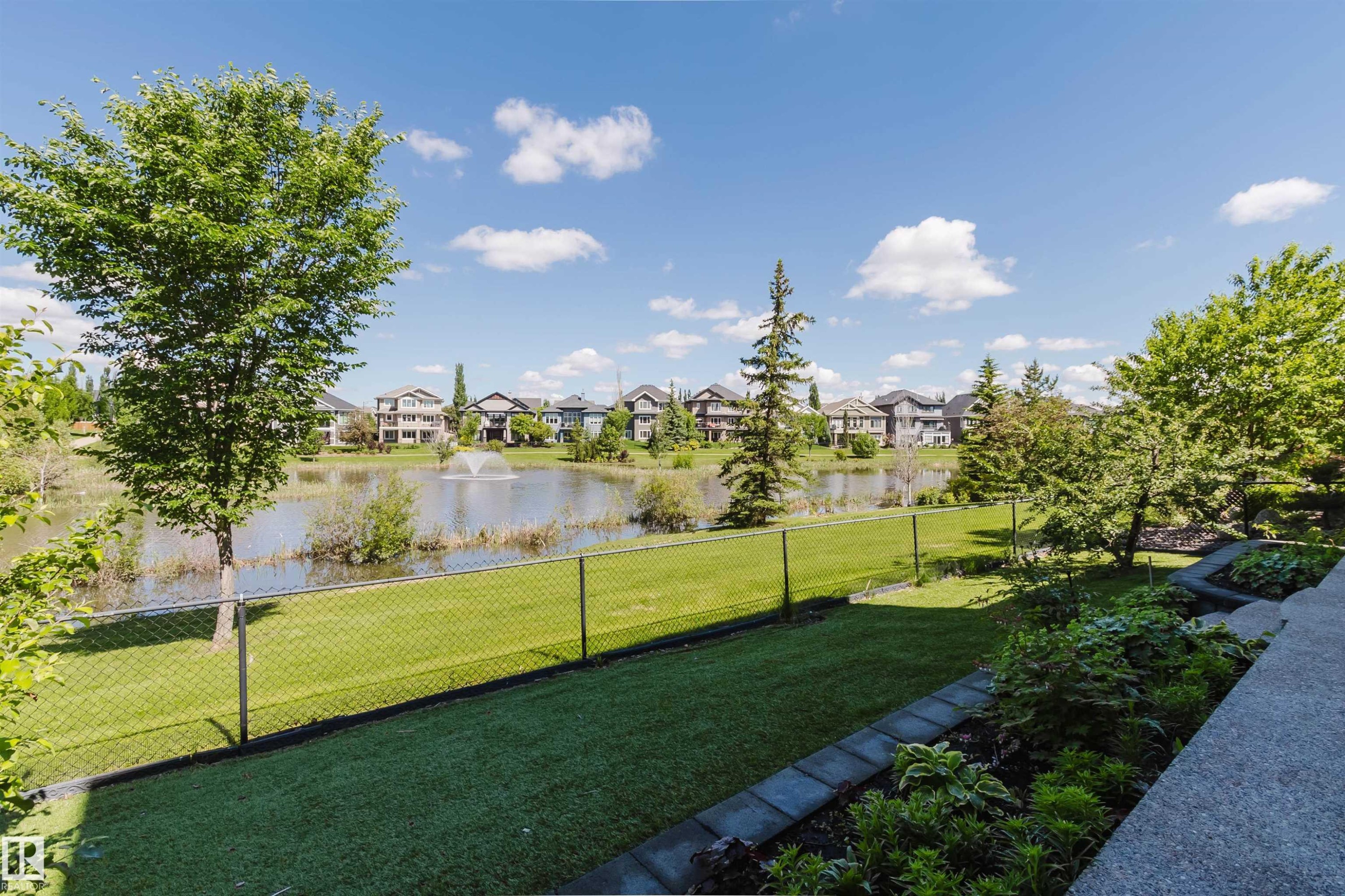 Landscaped backyard featuring a manicured lawn, established trees, and a chain-link fence bordering a pond with a fountain - 3438 West Landing Landing, Edmonton, AB