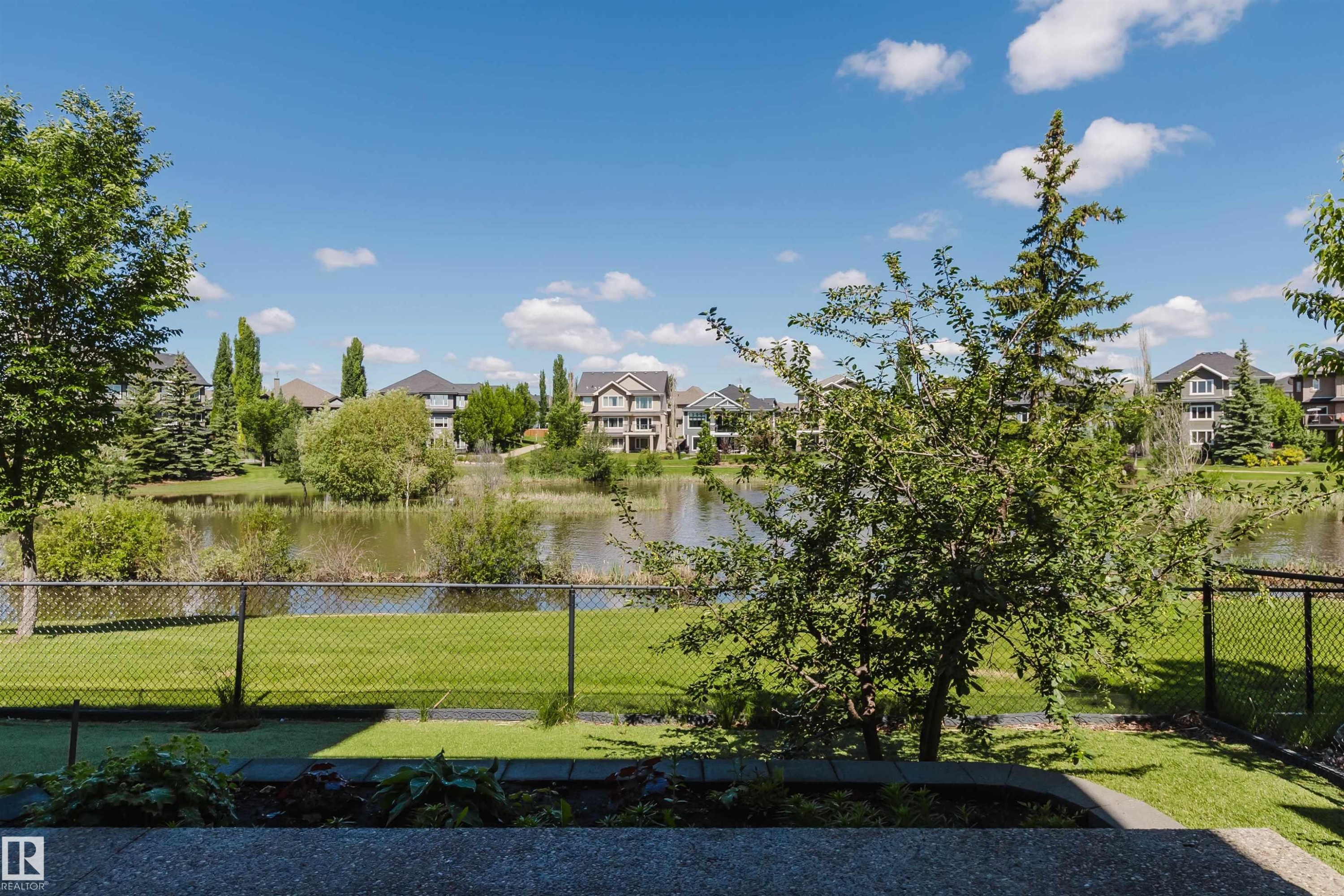 Landscaped backyard with chain-link fencing bordering a serene pond, expansive green space, and mature trees - 3438 West Landing Landing, Edmonton, AB