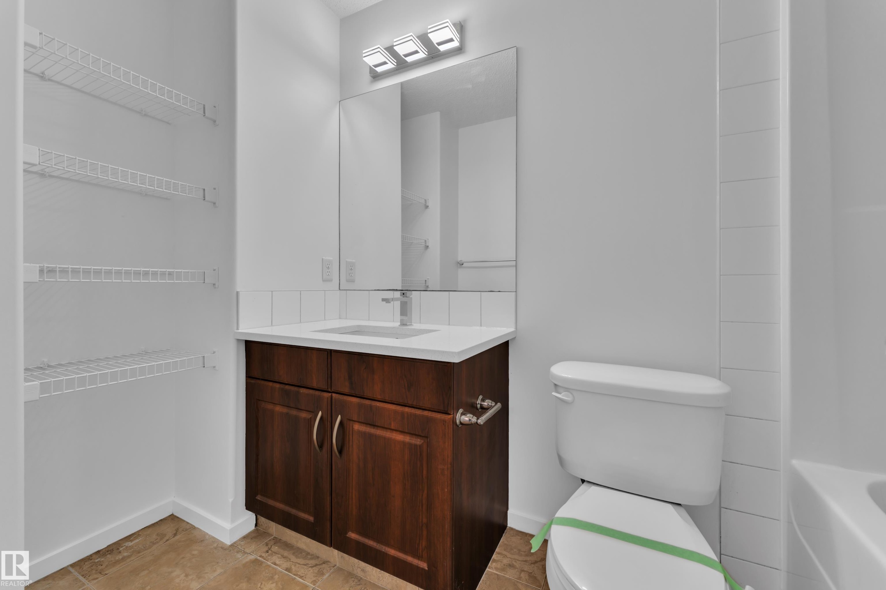 Contemporary bathroom featuring a dark wood-finish vanity with a white countertop and integrated sink, chrome faucet, and a rectangular mirror with a three-light fixture - 2143 32 Street, Edmonton, AB
