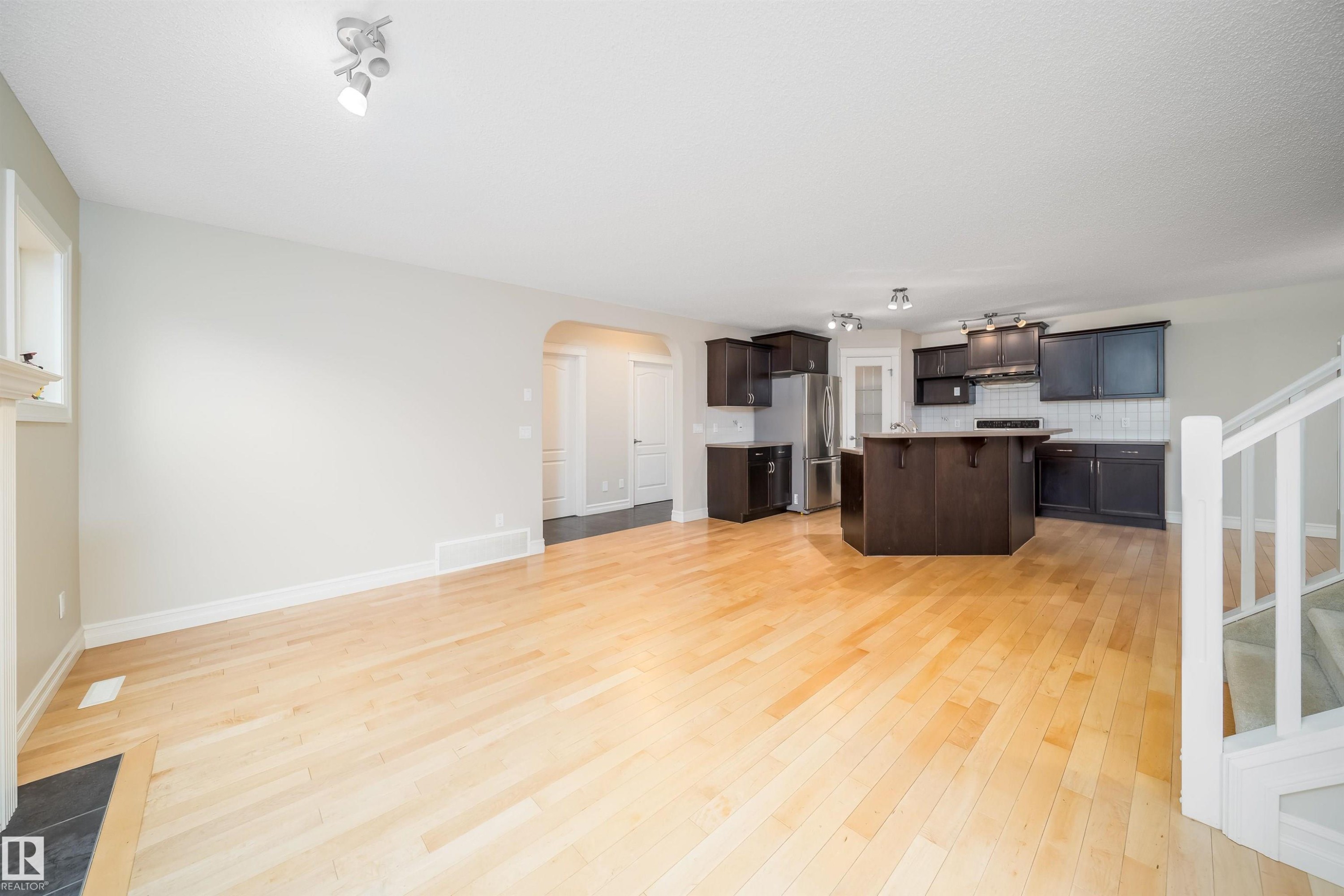Open-concept living area featuring light wood-finish flooring, a kitchen with dark cabinetry, stainless steel appliances, a center island with seating, and a white subway tile backsplash - 11312 13 Ave Sw, Edmonton, AB