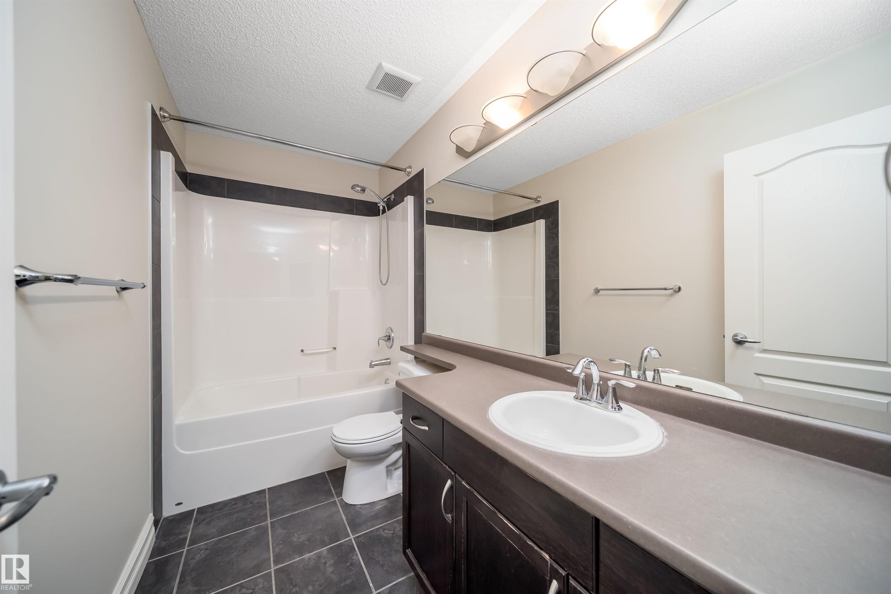 Bathroom featuring a white tub and shower combination with dark tile surround, a dark wood-finish vanity with an integrated sink, a light-toned countertop, and dark square floor tiles - 11312 13 Ave Sw, Edmonton, AB