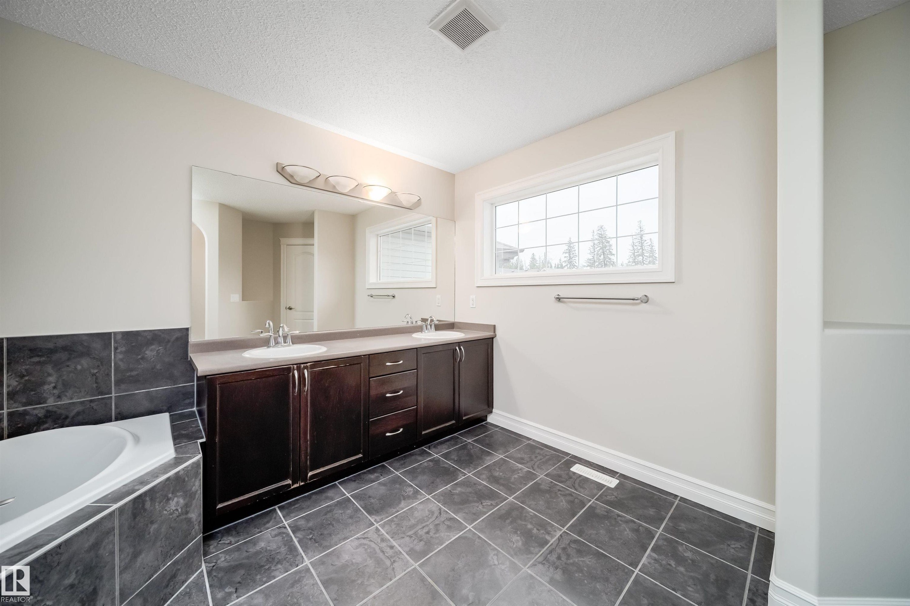 Full-size bathroom featuring a double vanity with ample storage, a large soaking tub with dark tile surround, a spacious window, and dark tiled flooring - 11312 13 Ave Sw, Edmonton, AB