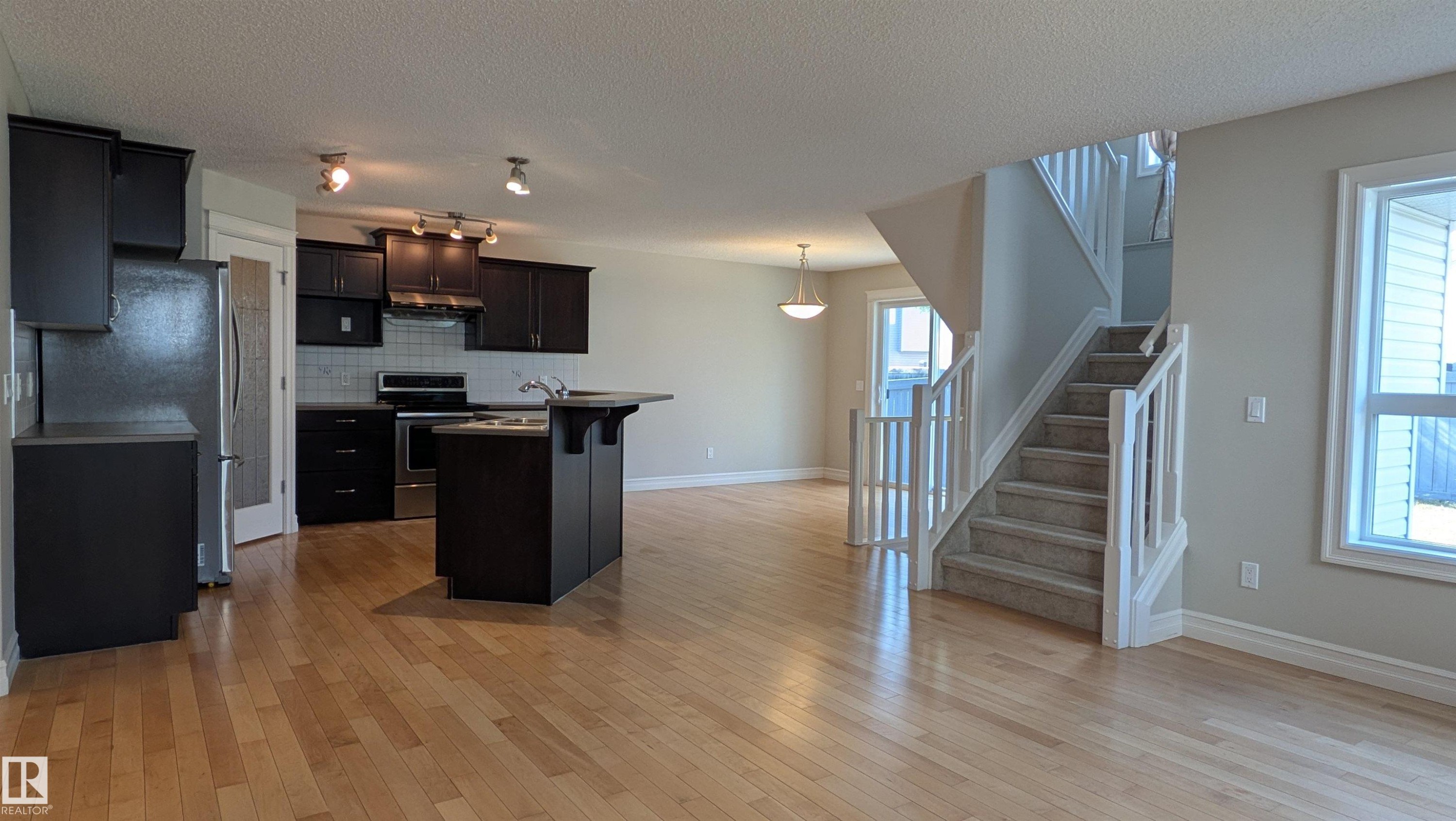 Open-concept living area featuring wood-finish flooring, a kitchen with dark cabinetry and stainless steel appliances, a central kitchen island with a sink, and a carpeted staircase with white railings - 11312 13 Ave Sw, Edmonton, AB