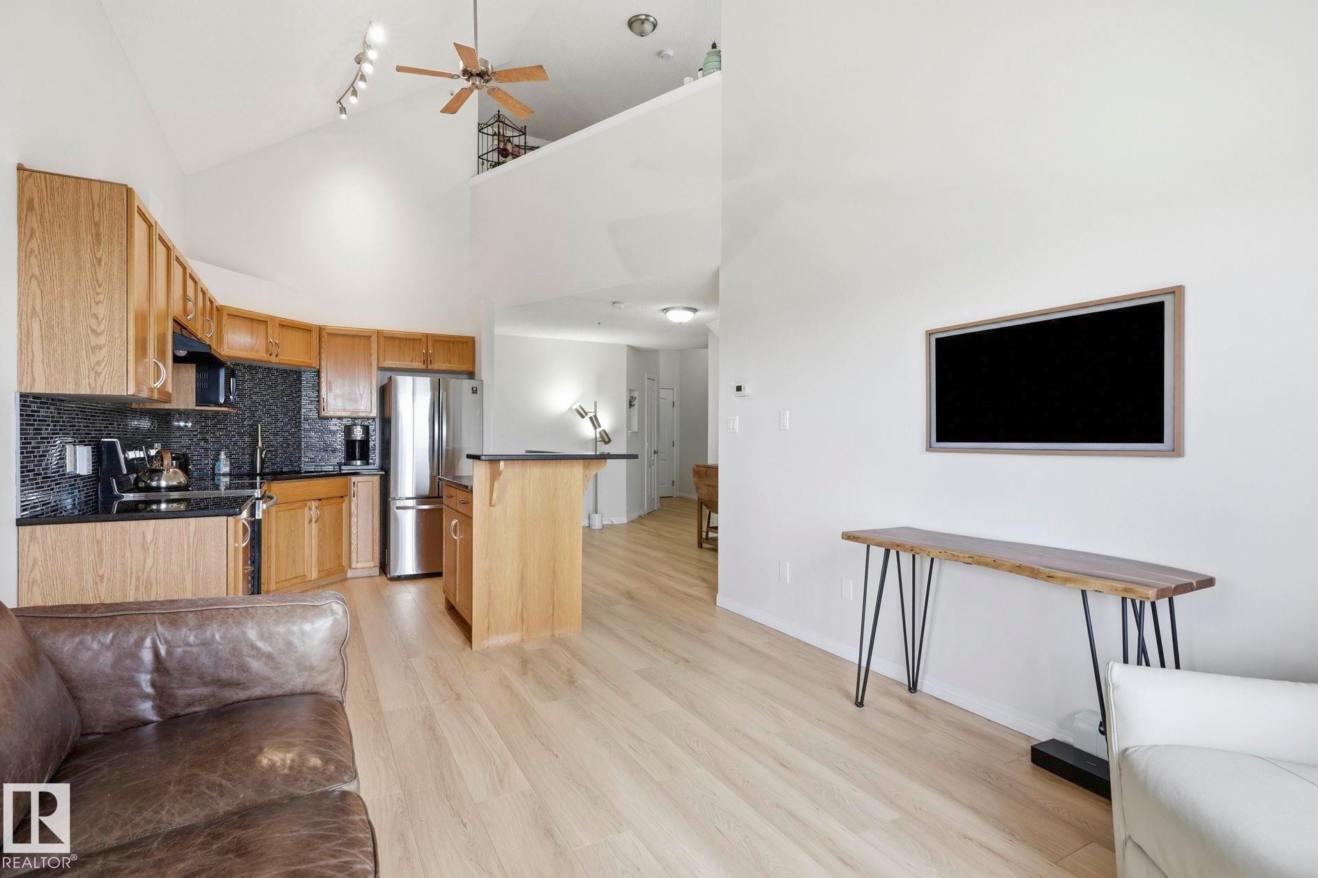 Open-concept living area featuring vaulted ceilings, light wood-finish flooring, a kitchen with wood cabinetry and dark countertops, and a mezzanine balcony overhead - #426 3425 19 St Nw, Edmonton, AB