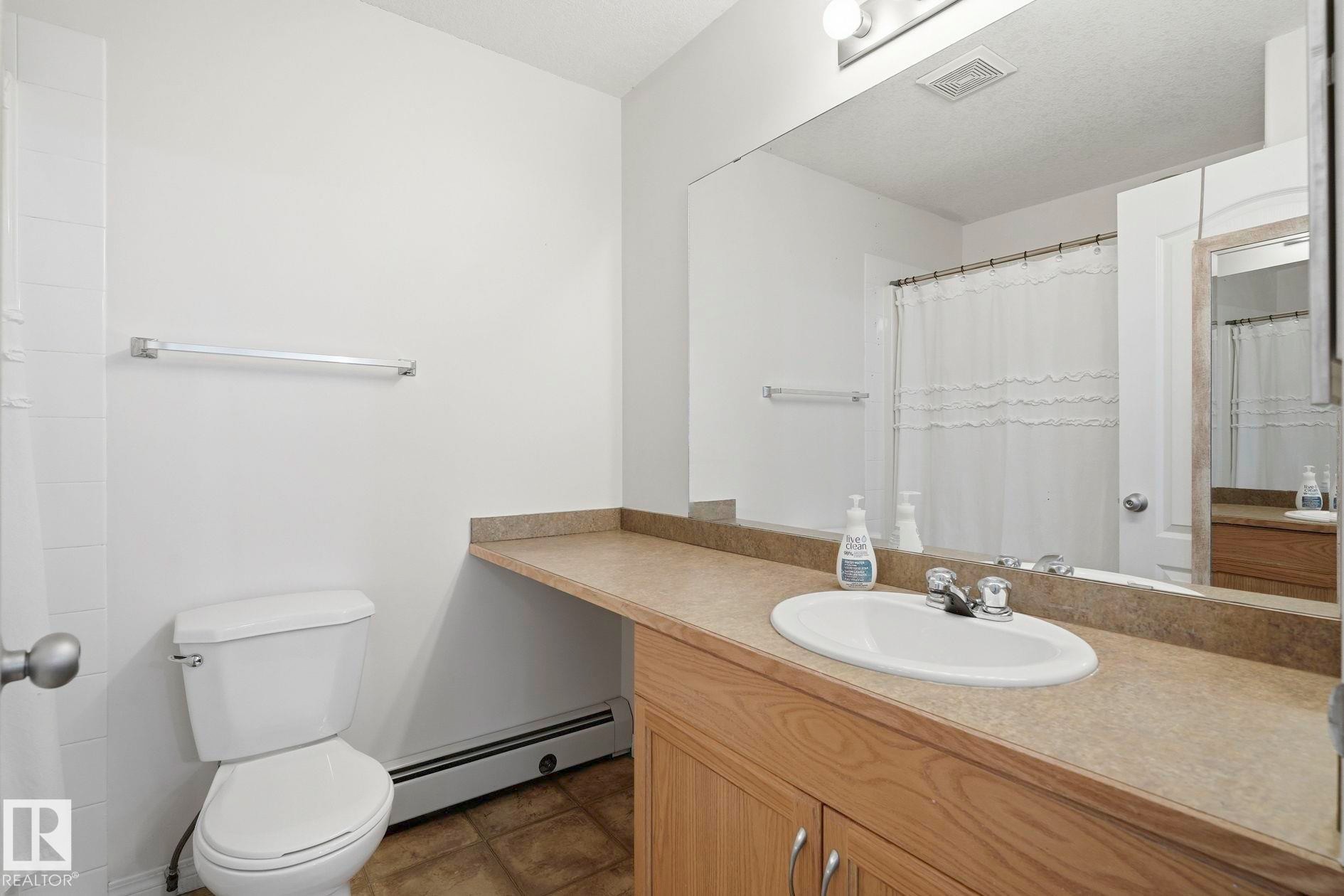 Bathroom featuring a wood-finish vanity with an integrated sink, a full-width mirror, and a white toilet - #426 3425 19 St Nw, Edmonton, AB