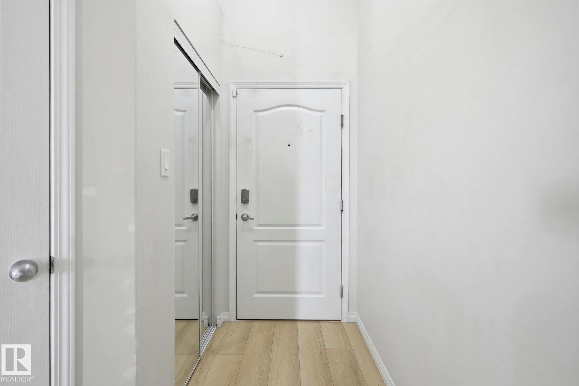 Entryway featuring light wood-finish flooring, a white paneled door, and a mirrored closet with sliding doors - #426 3425 19 St Nw, Edmonton, AB