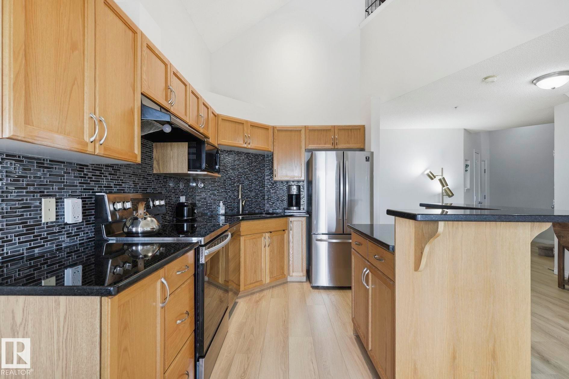 Kitchen featuring light wood cabinetry, dark mosaic tile backsplash, black countertops, stainless steel appliances, and wood-finish flooring - #426 3425 19 St Nw, Edmonton, AB