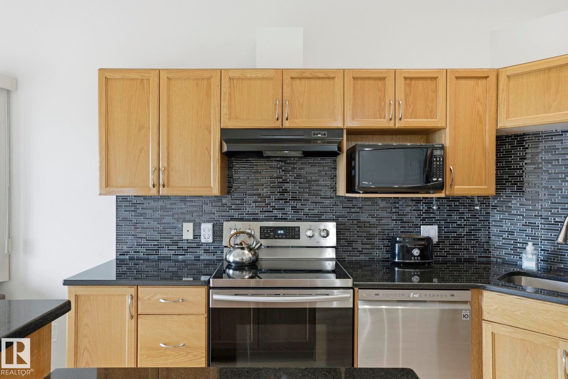 Kitchen featuring light wood cabinetry, dark countertops, a mosaic tile backsplash, and stainless steel appliances including a range and dishwasher - #426 3425 19 St Nw, Edmonton, AB