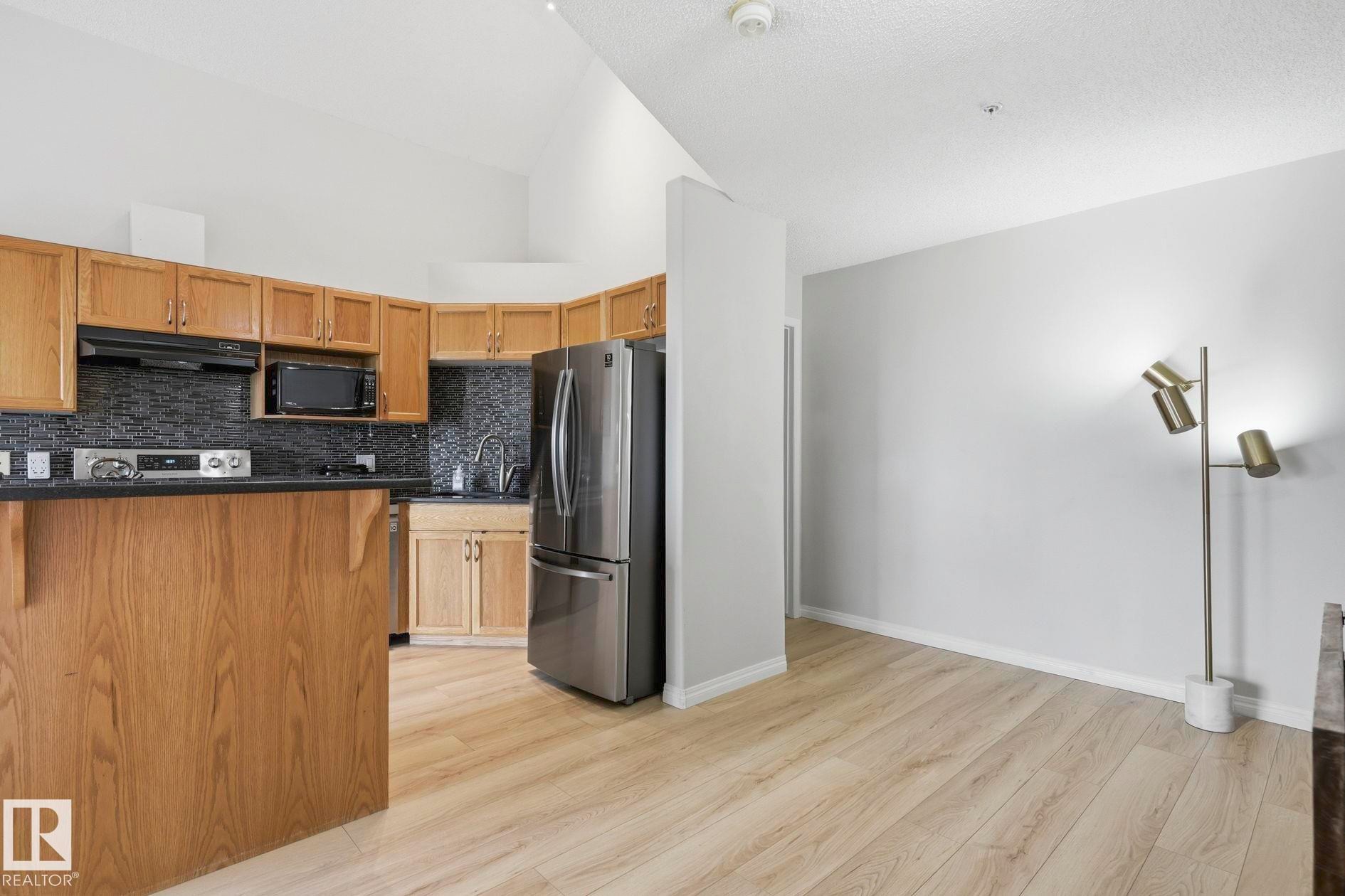 Open-concept kitchen featuring light wood cabinetry, dark countertops, dark mosaic tile backsplash, and stainless steel appliances, all complemented by wood-finish flooring and a vaulted ceiling - #426 3425 19 St Nw, Edmonton, AB