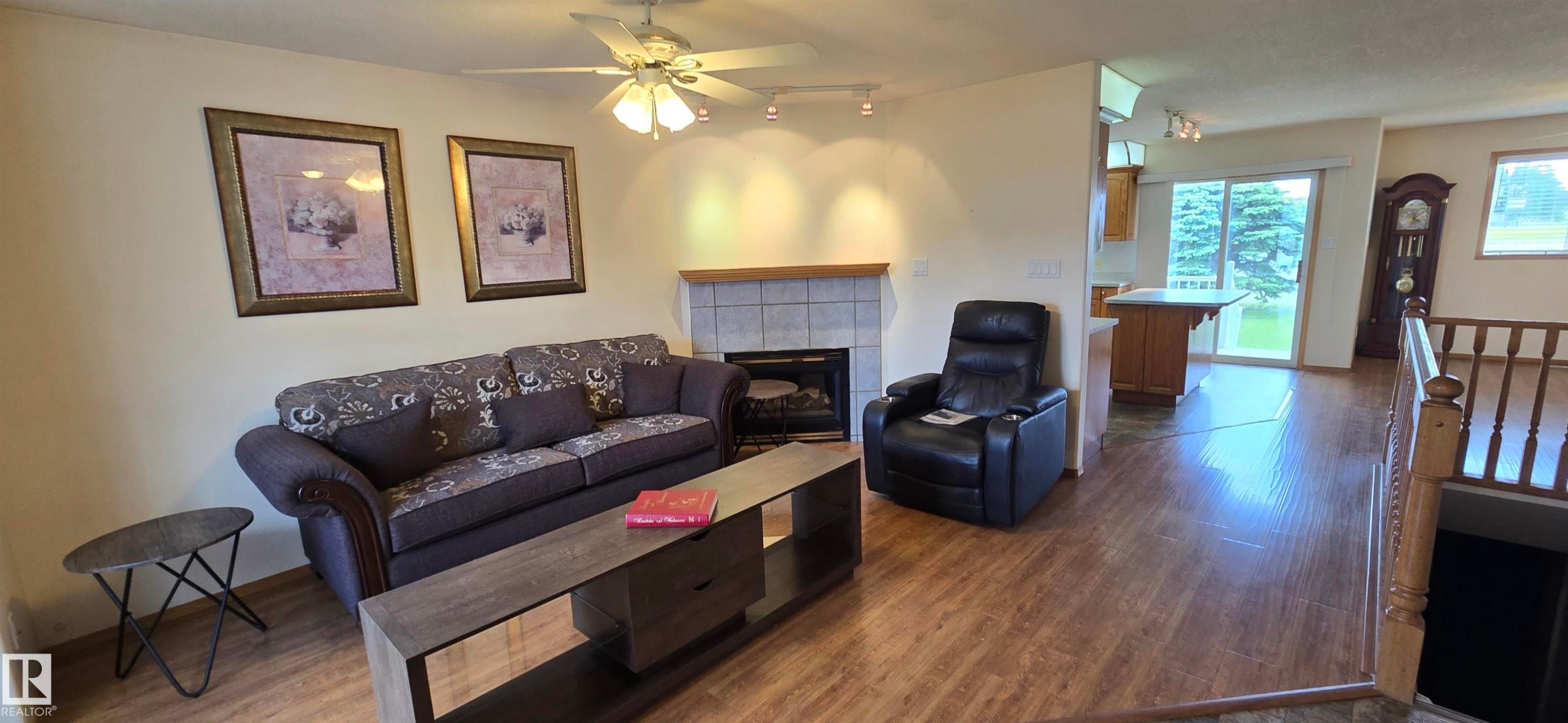 Living area featuring wood-finish flooring, a ceiling fan with integrated lighting, and a tiled fireplace with a mantel - 4519 59 Ave, Vegreville, AB