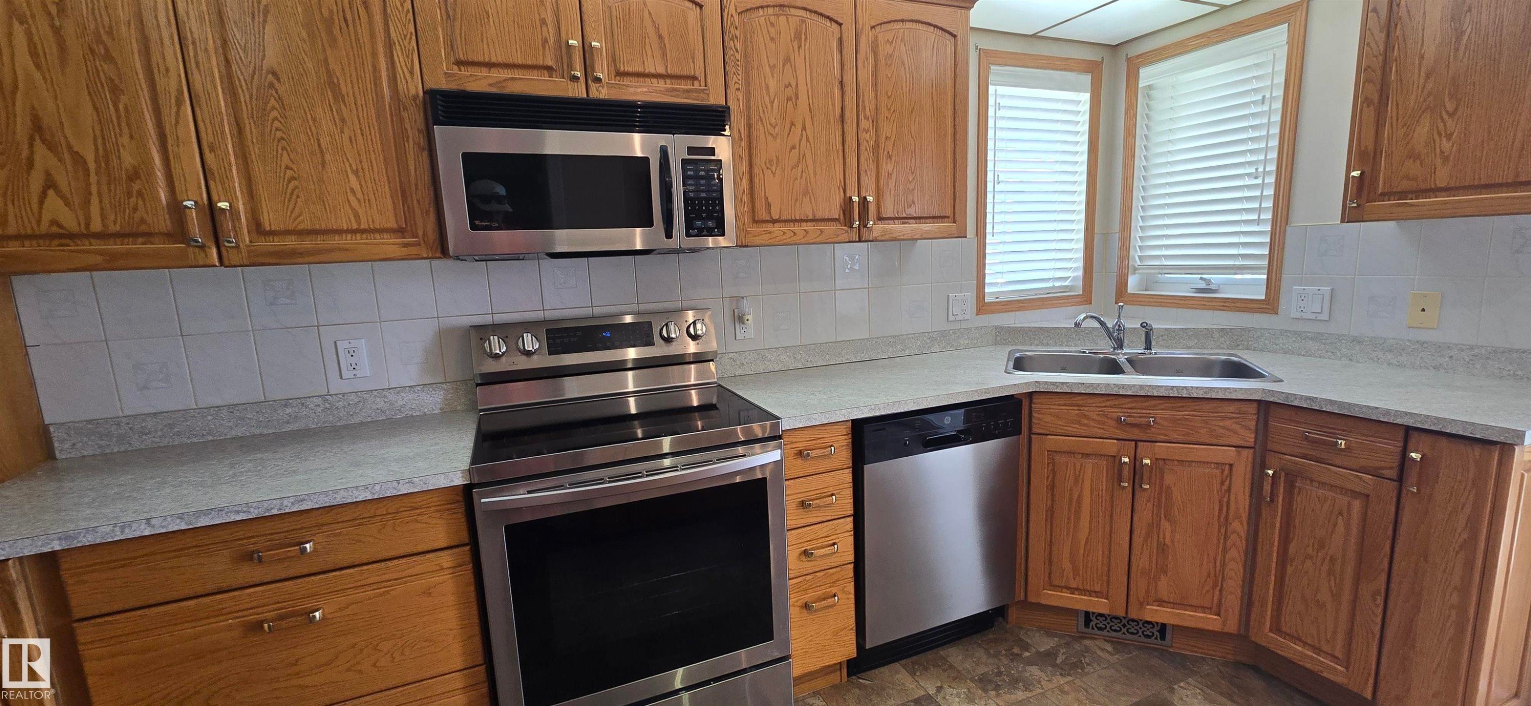 Kitchen featuring wood cabinetry, stainless steel appliances, white subway tile backsplash, light-toned countertops, and a double basin sink - 4519 59 Ave, Vegreville, AB