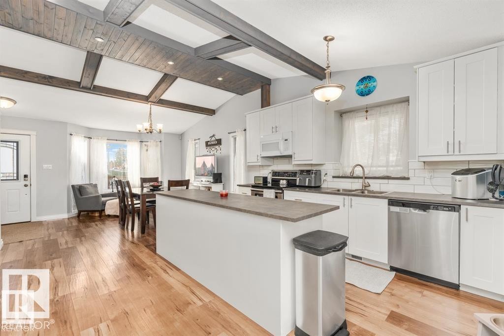 Open-concept living space with vaulted ceilings featuring exposed wood beams, wood-finish flooring, a kitchen island with a solid surface countertop, white cabinetry, and stainless steel appliances - 1112 13 Avenue, Cold Lake, AB - Indoor Photo Showing Kitchen With Double Sink With Upgraded Kitchen