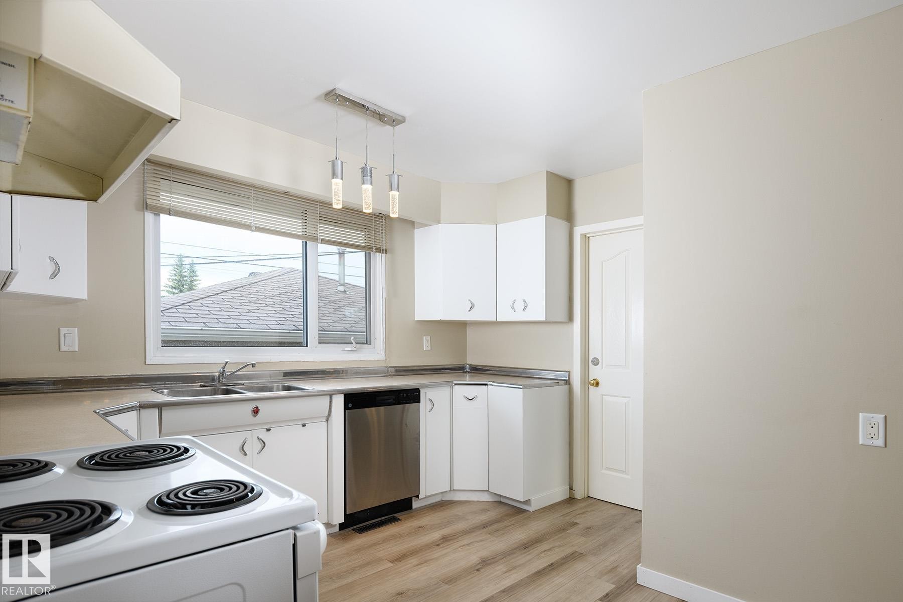 Kitchen featuring white cabinetry, wood-finish flooring, stainless steel dishwasher, double basin sink, and a multi-light pendant fixture - 9607 75 Street, Edmonton, AB