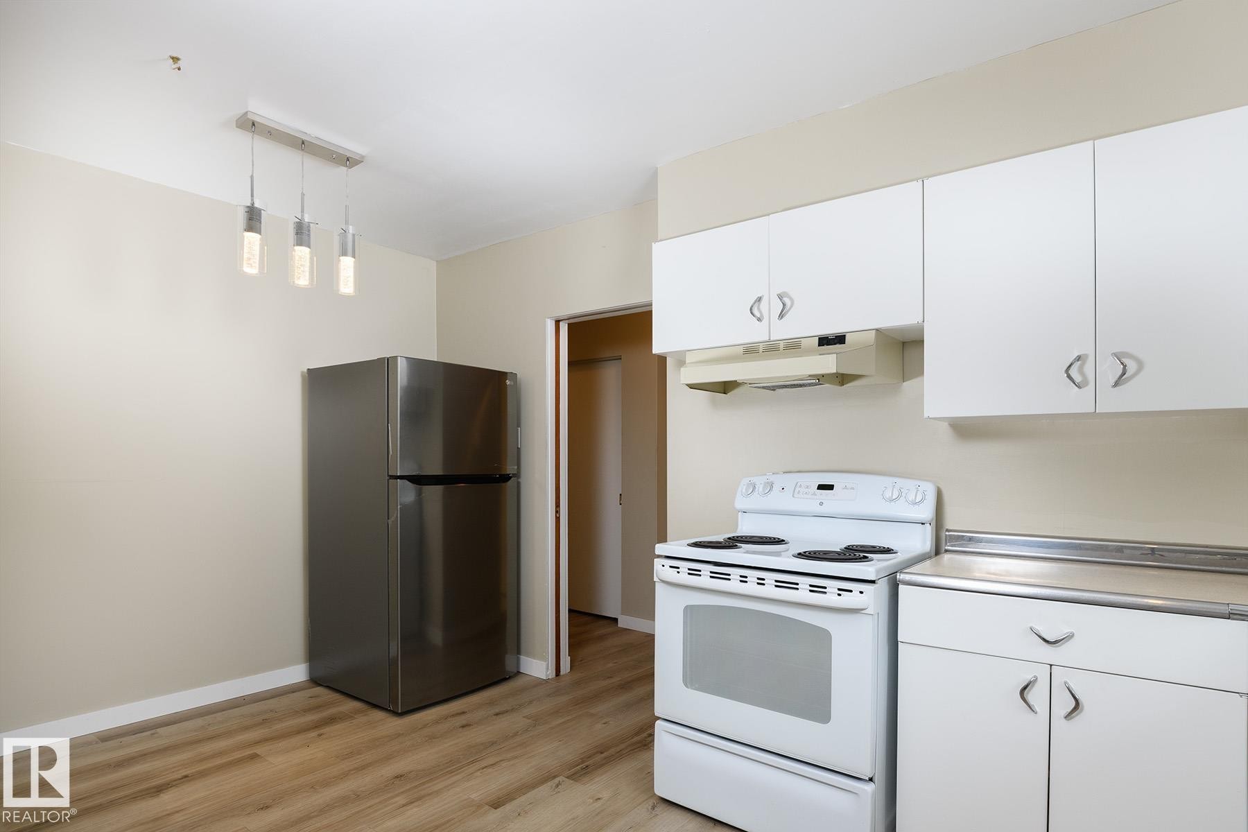Kitchen space featuring wood-finish flooring, a stainless steel refrigerator, white cabinetry with silver hardware, a white electric range, and a brushed nickel linear pendant light fixture - 9607 75 Street, Edmonton, AB